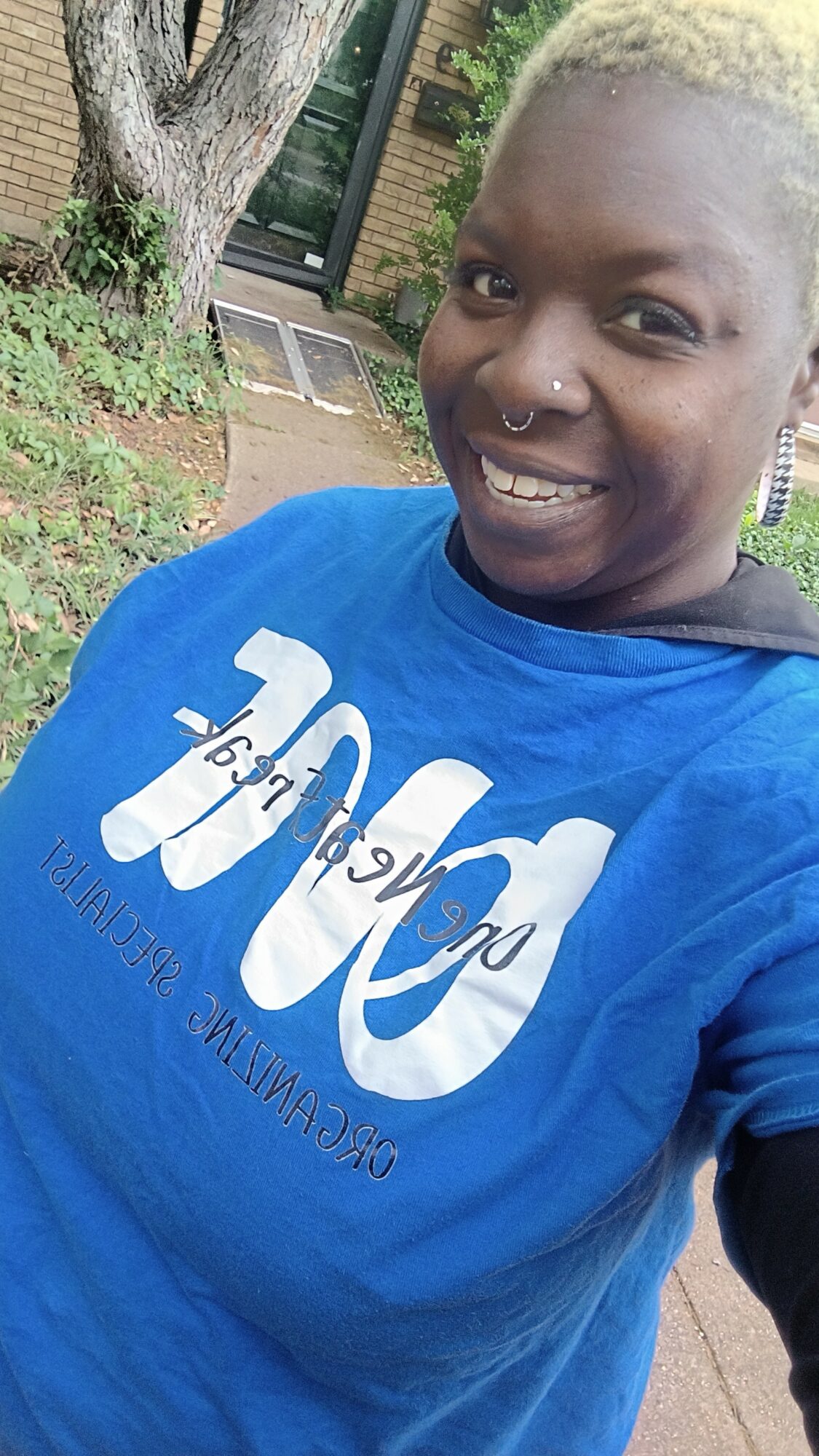 Person with short blonde hair smiling outdoors, wearing a blue shirt with white text, standing near a brick building and tree.