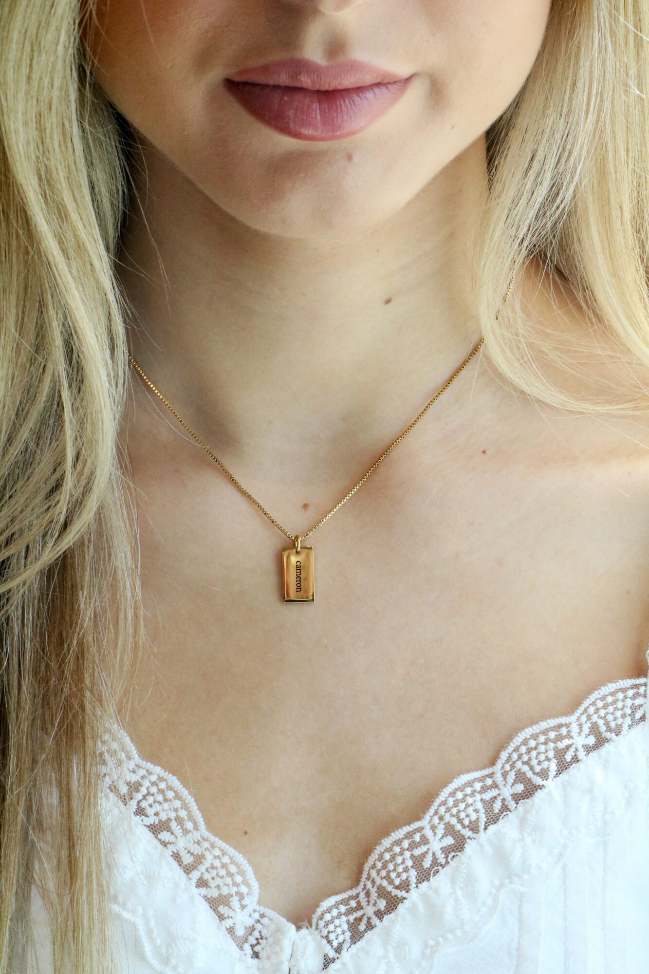 Close-up of a woman wearing a gold necklace with a rectangular pendant, white lace top, and light blonde hair.