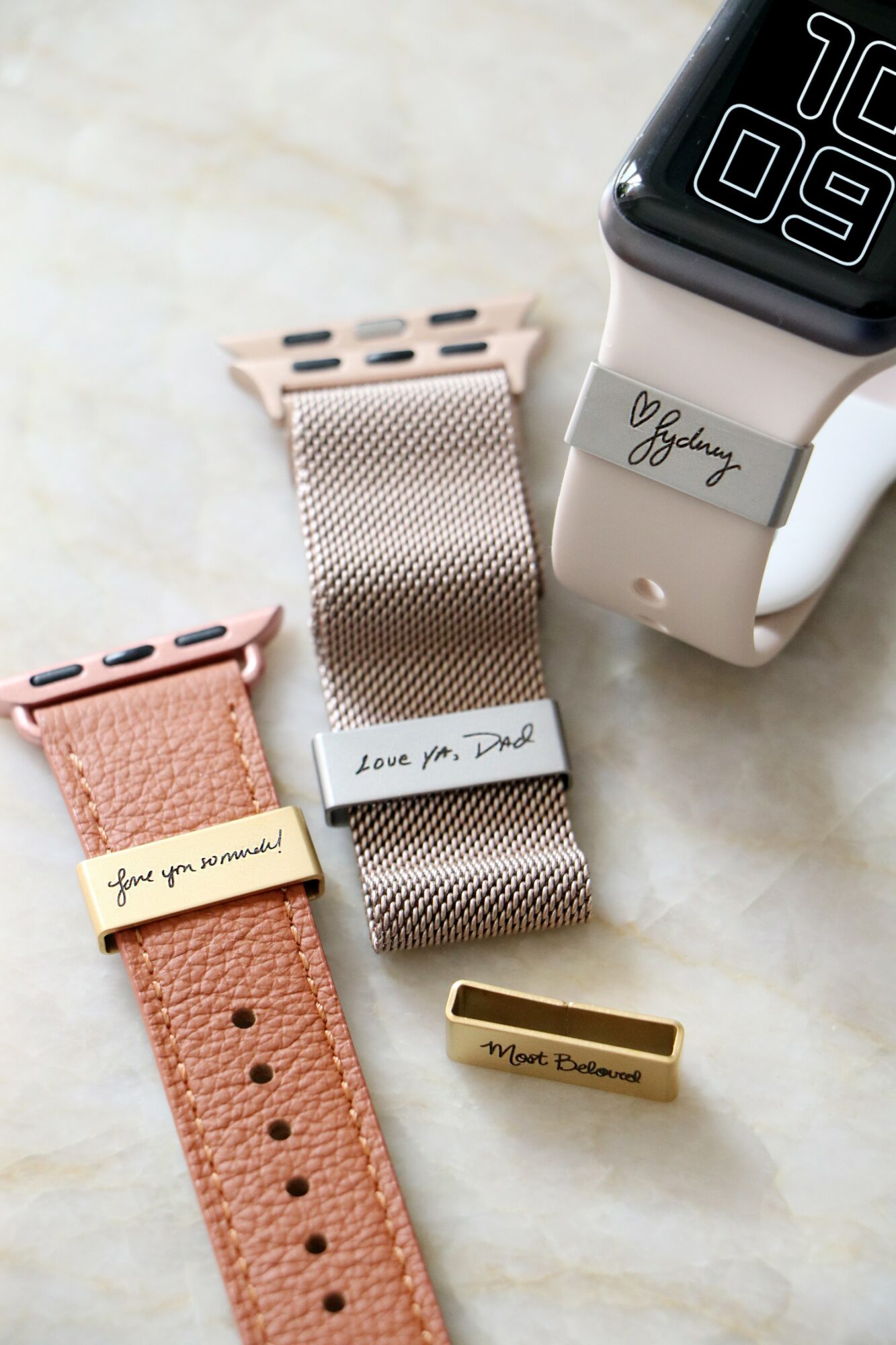 Two smartwatch bands, one pink and one silver, with handwritten notes and a smartwatch displaying a digital clock, on a light surface.