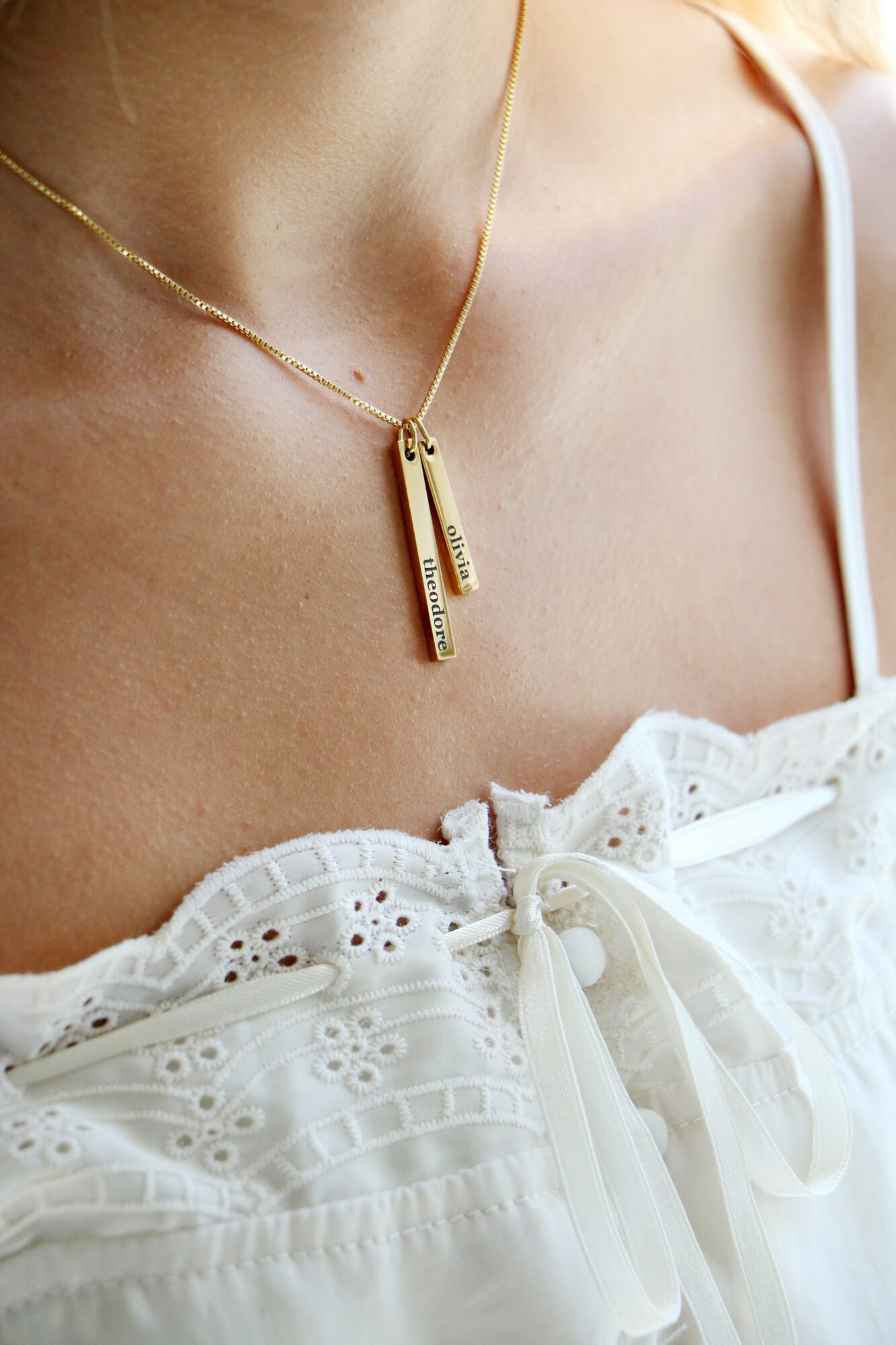 Close-up of a woman wearing a gold necklace with two vertical bars and a white eyelet dress with a bow detail.