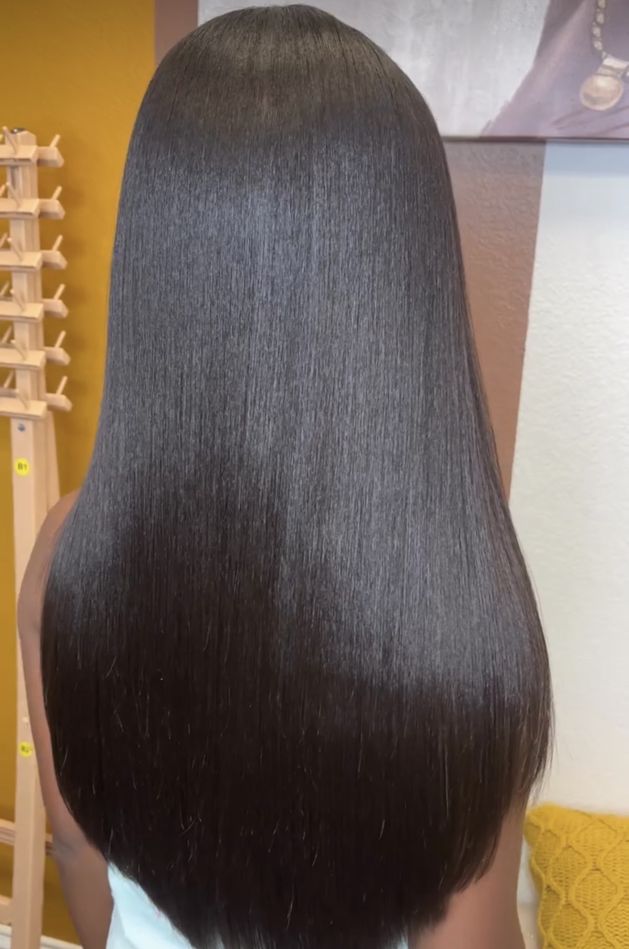 Back of a person with long, straight, shiny black hair, indoors with yellow and purple decor visible