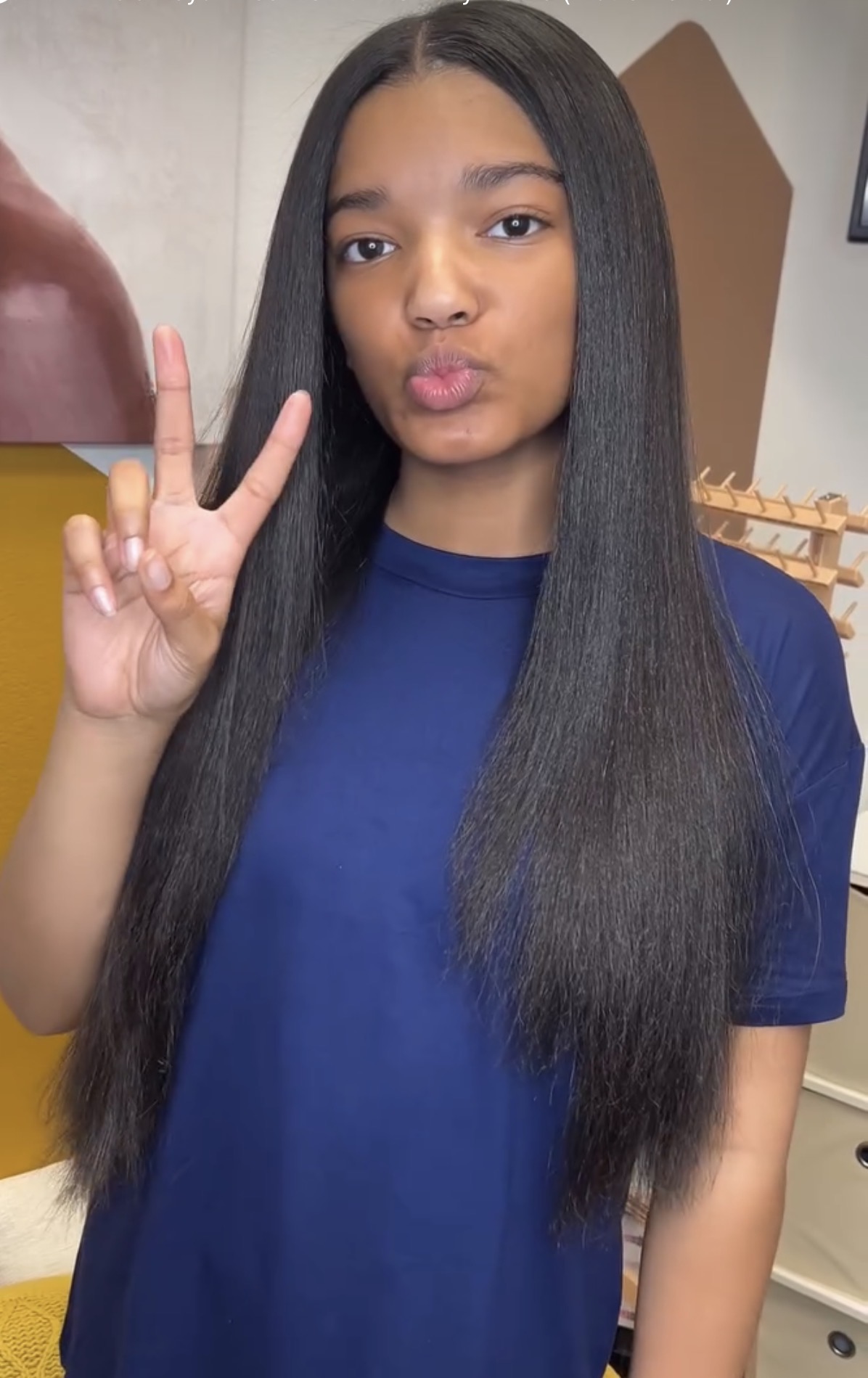 Young woman with long, straight black hair making a peace sign gesture, wearing a blue shirt, indoors.