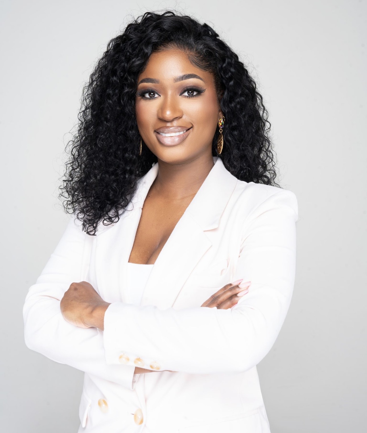 Woman with curly hair in a white blazer smiling with arms crossed, neutral background.
