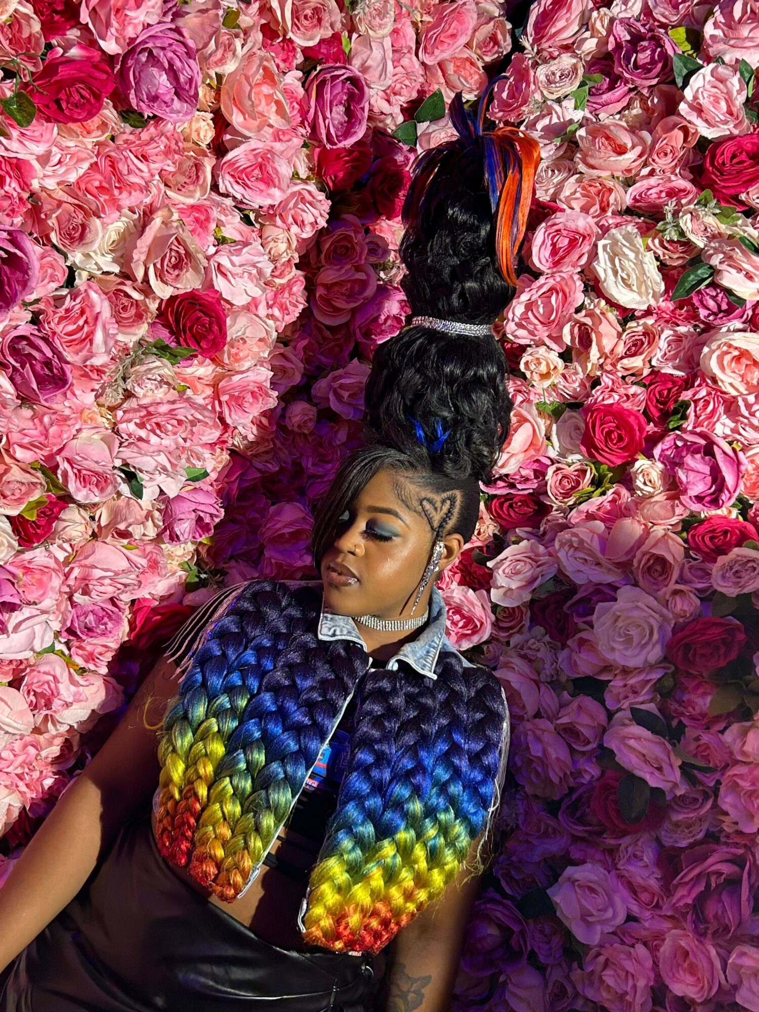 Person lying on a bed of pink and purple roses, wearing a colorful, rainbow-patterned vest and a black outfit, with hair styled in a high bun.