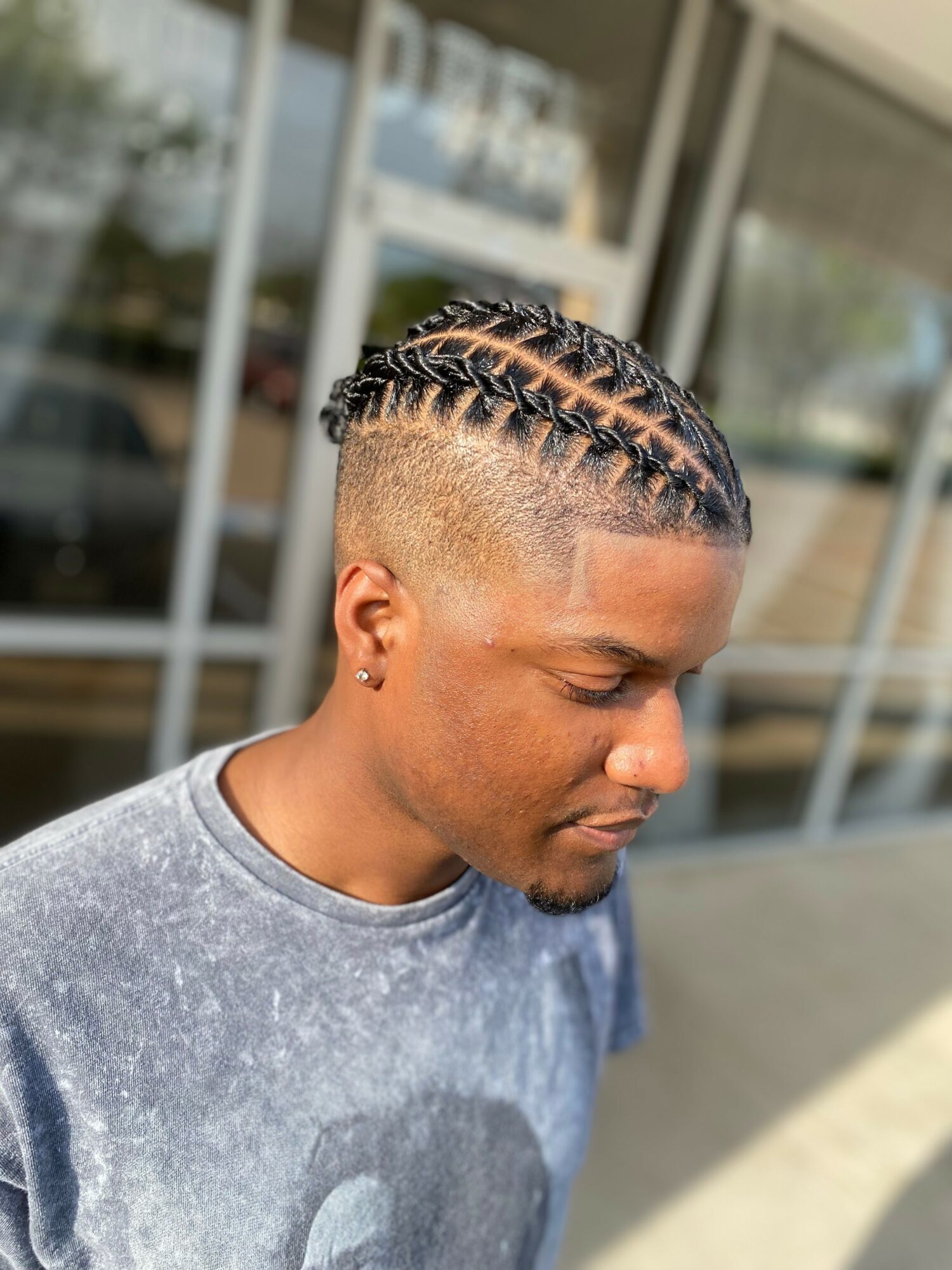 Young man with braided hairstyle looking downward outside a building, wearing a gray T-shirt.