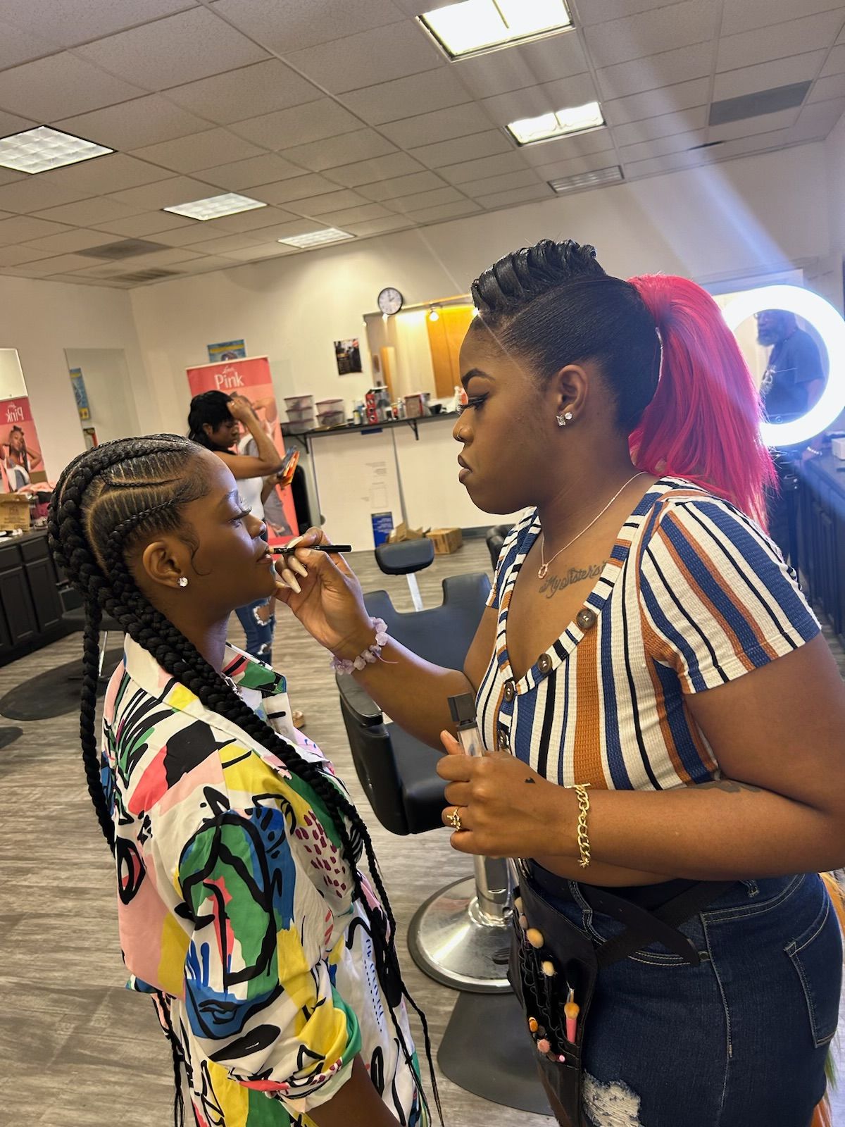 Two women with braided hair and colorful clothing are talking in a salon, with hair styling tools and mirrors visible.