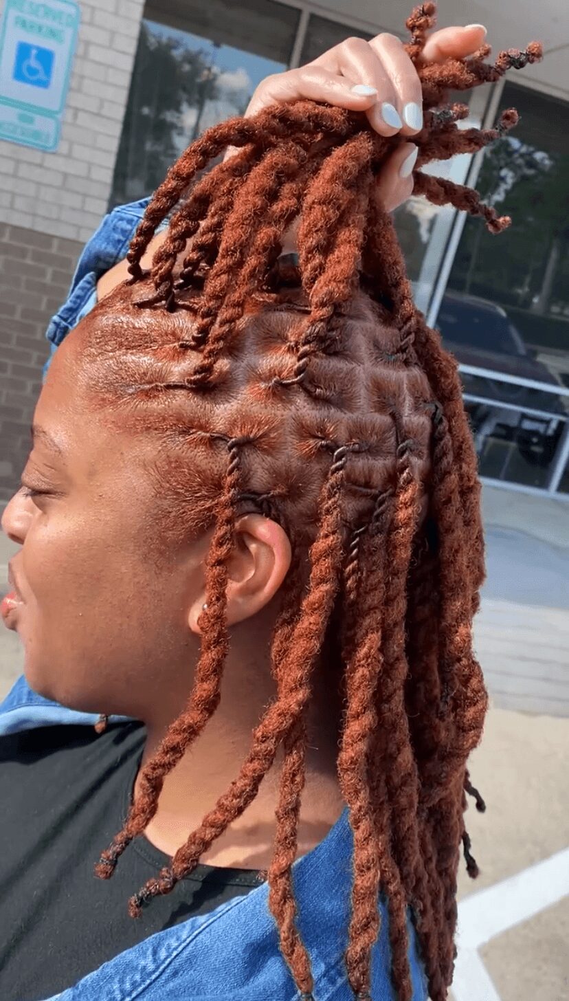 Person with styled dreadlocks, holding up hair outside a building with glass windows and a parking lot.