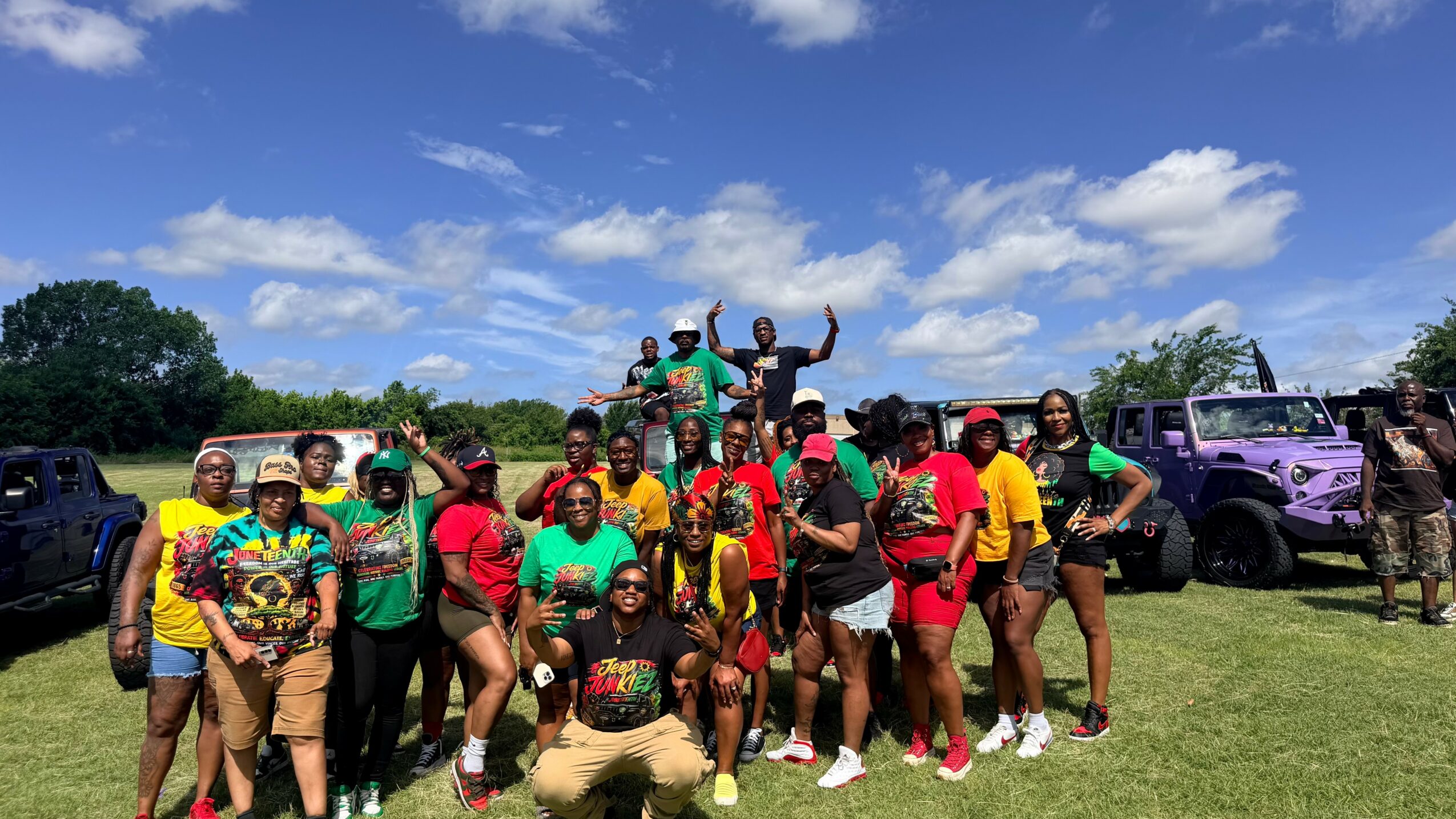 Group of people outdoors on grass, some raising hands, with cars and trees in background, under blue sky with clouds.