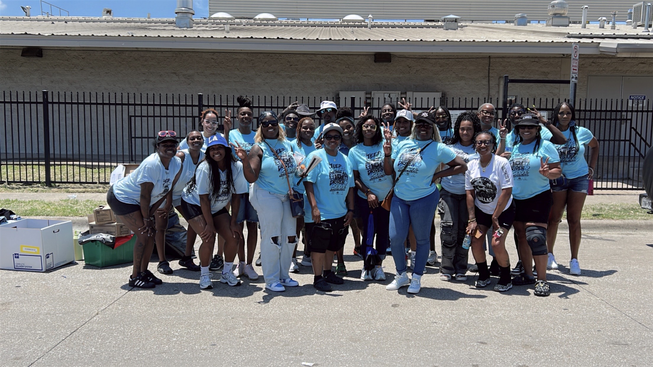 Group of people standing outdoors in front of a fence and building, some wearing matching light blue shirts, posing for a photo.