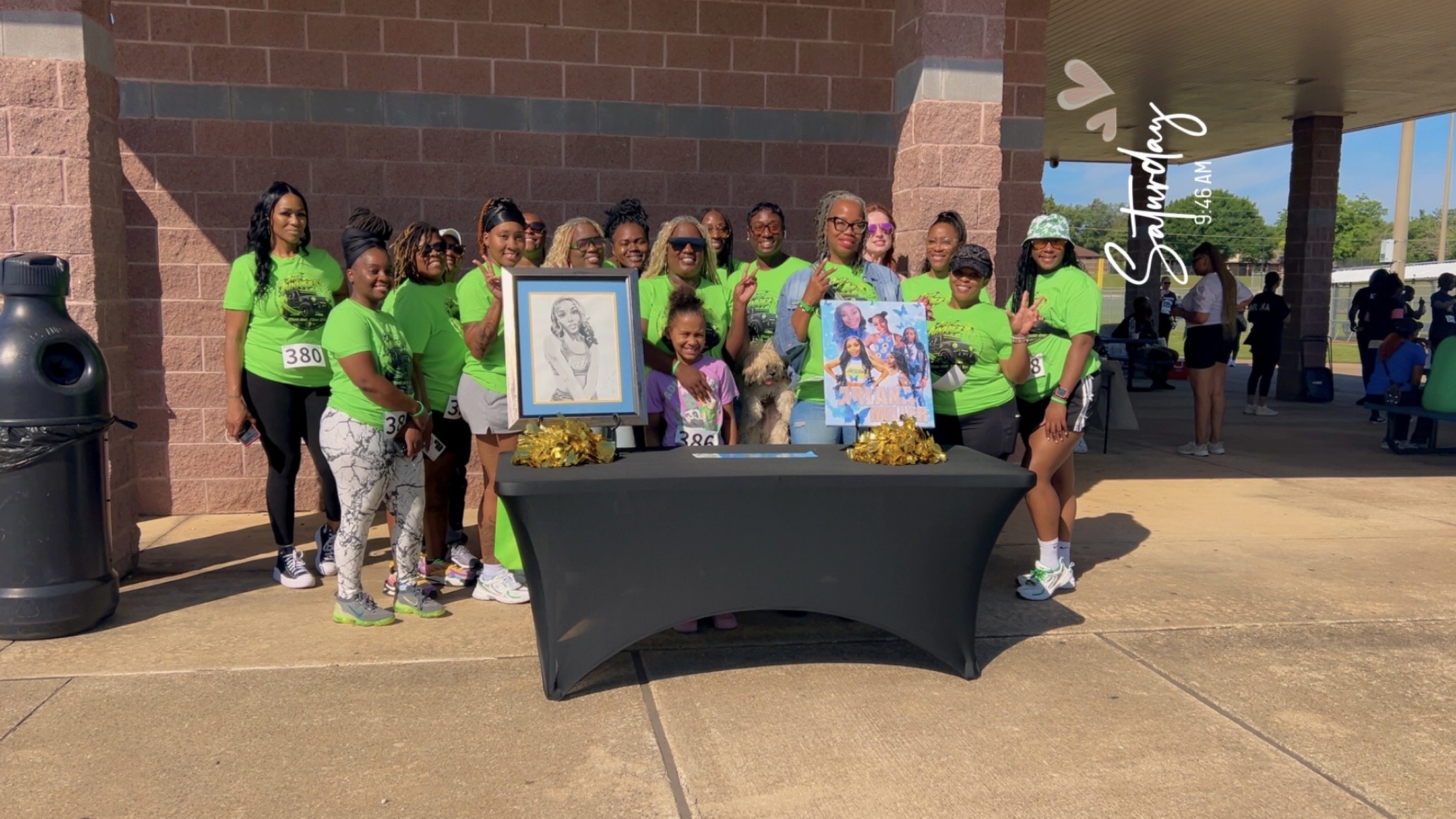 Group of people in green shirts gathered behind a table with artwork and decorations outside a building.