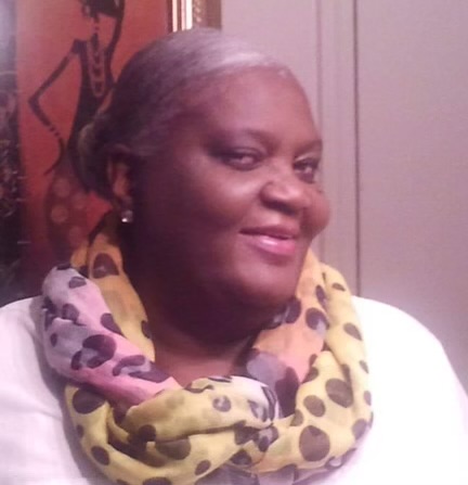 Smiling woman with gray hair tied back, wearing a yellow and purple polka dot scarf, white top, and earrings.