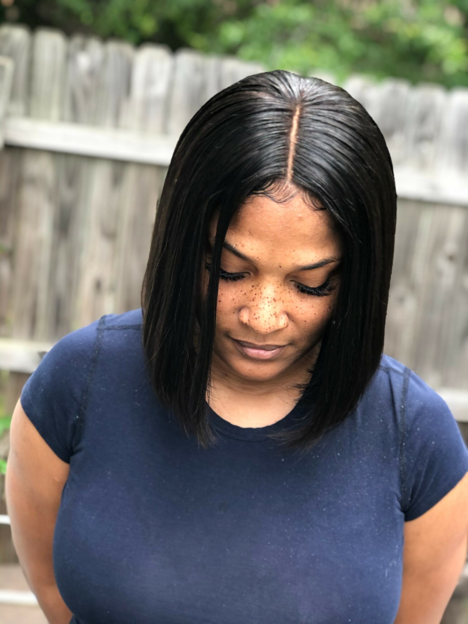 Woman with straight black hair wearing a navy blue shirt outdoors, looking down, wooden fence and greenery in background.