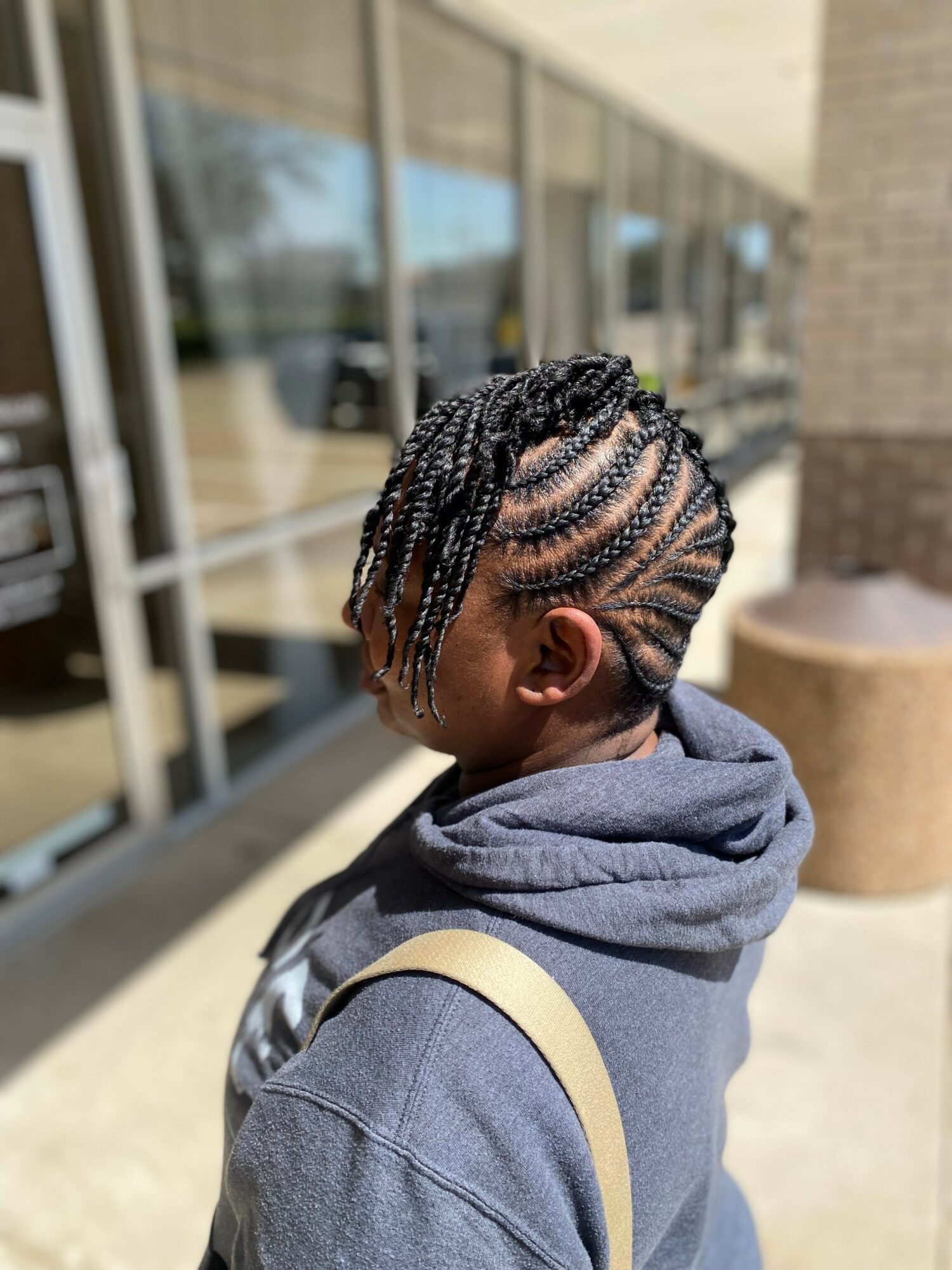Side view of a person with braided hairstyle, wearing a hoodie and backpack, outside a building with glass windows.