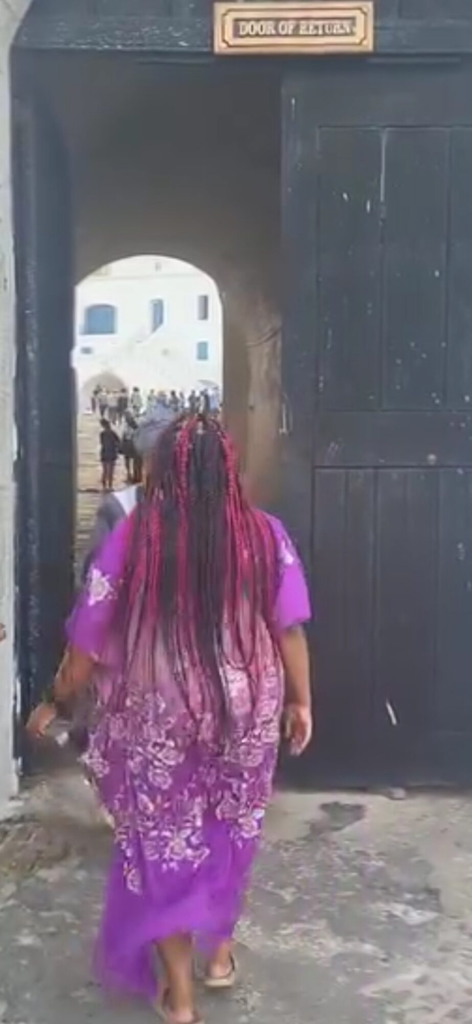 Person with long pink hair wearing a purple dress walking through a black gate, with a white building in the background.