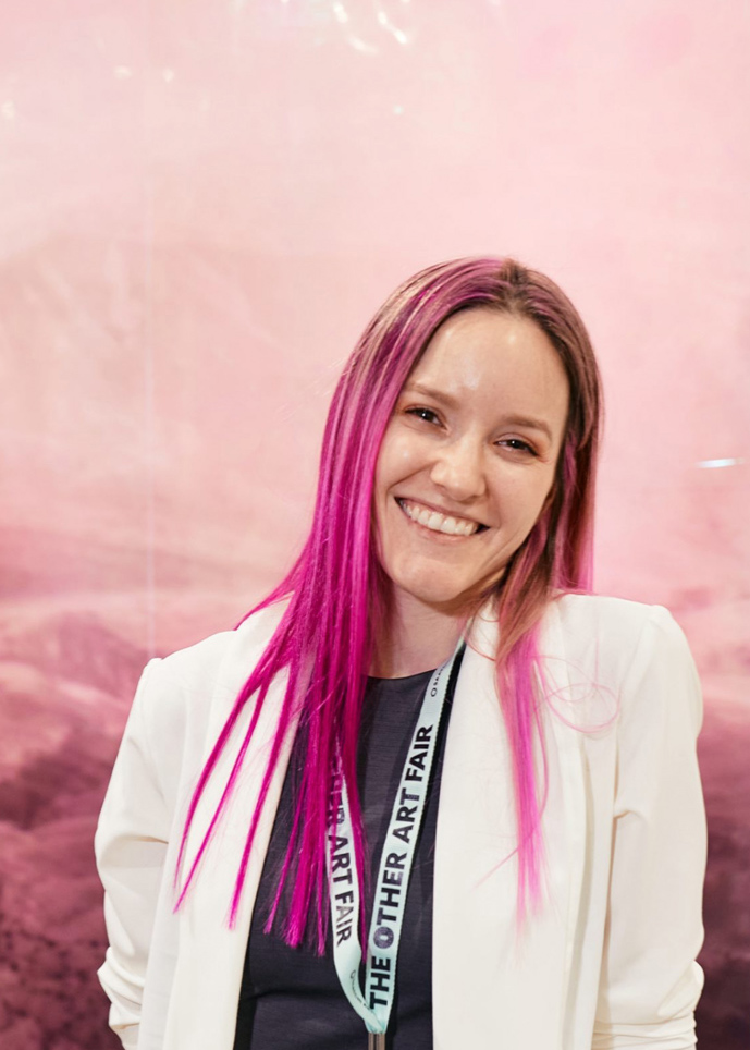 Young woman with long pink hair smiling, wearing a white blazer and a lanyard, against a pink background.