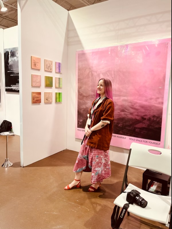 Woman standing in an art gallery with colorful paintings and a large pink abstract artwork behind her.
