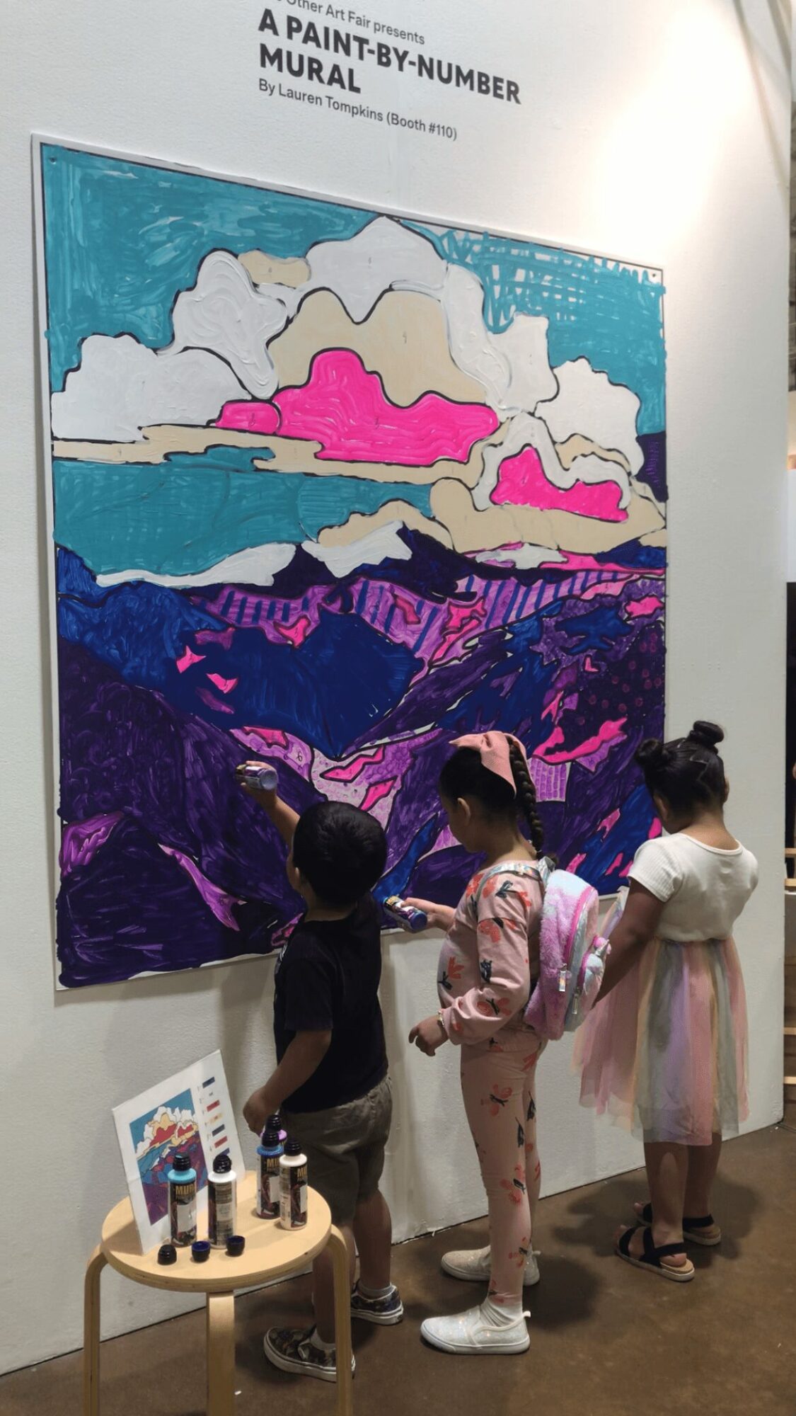 Three children stand in front of a colorful mural, observing the artwork on the wall. A small table with art supplies is nearby.