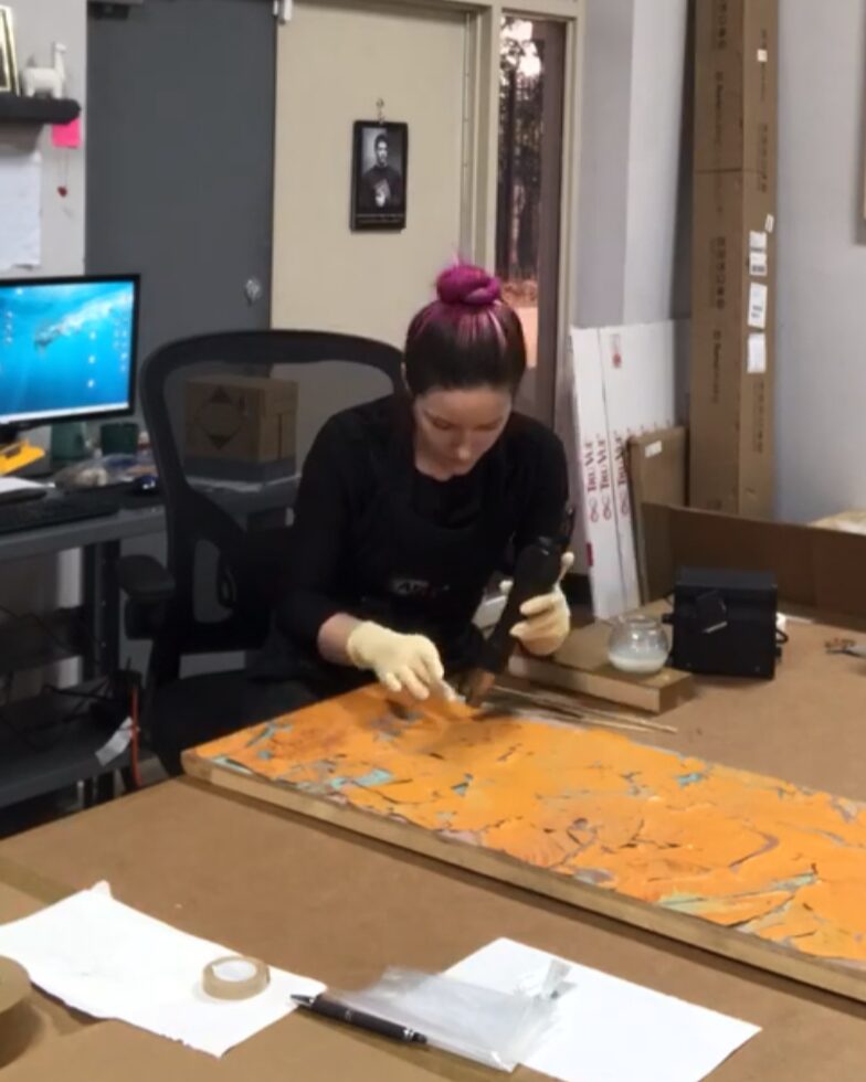 Person with pink hair tied in a bun working on a long orange and yellow artwork at a table, wearing gloves.
