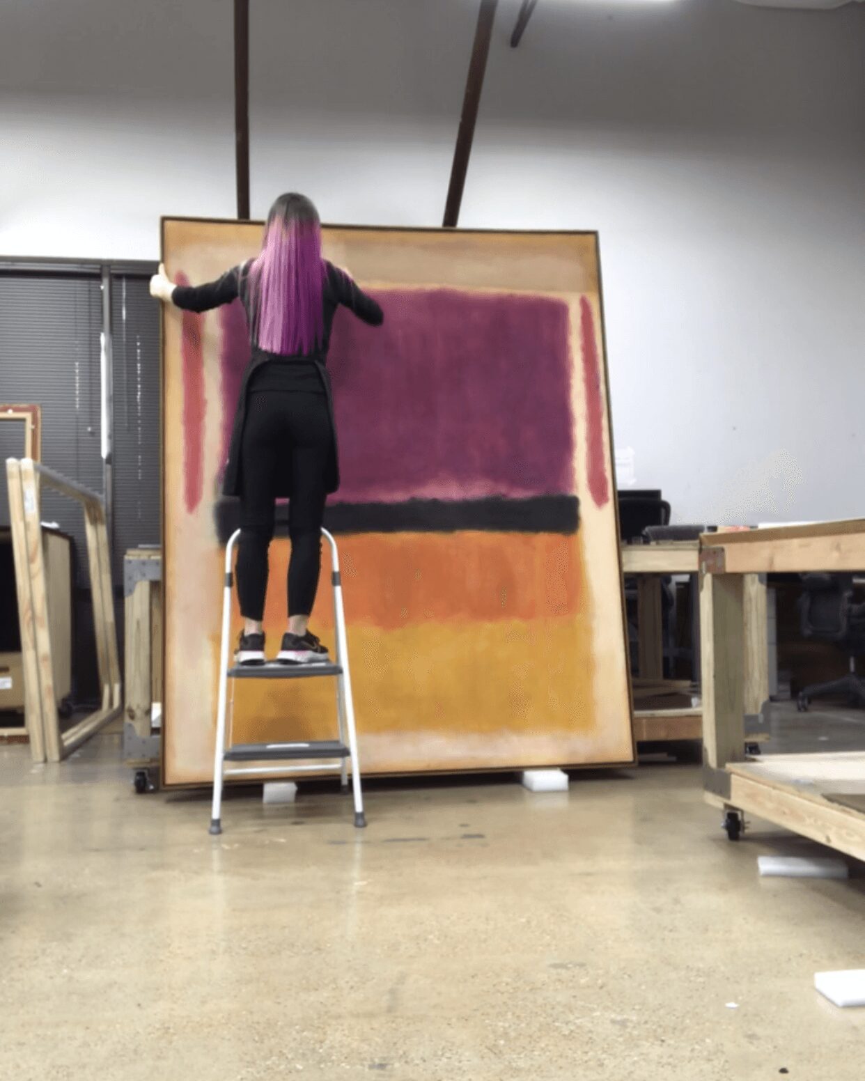 Person with purple hair painting on large canvas in workshop, standing on a ladder.
