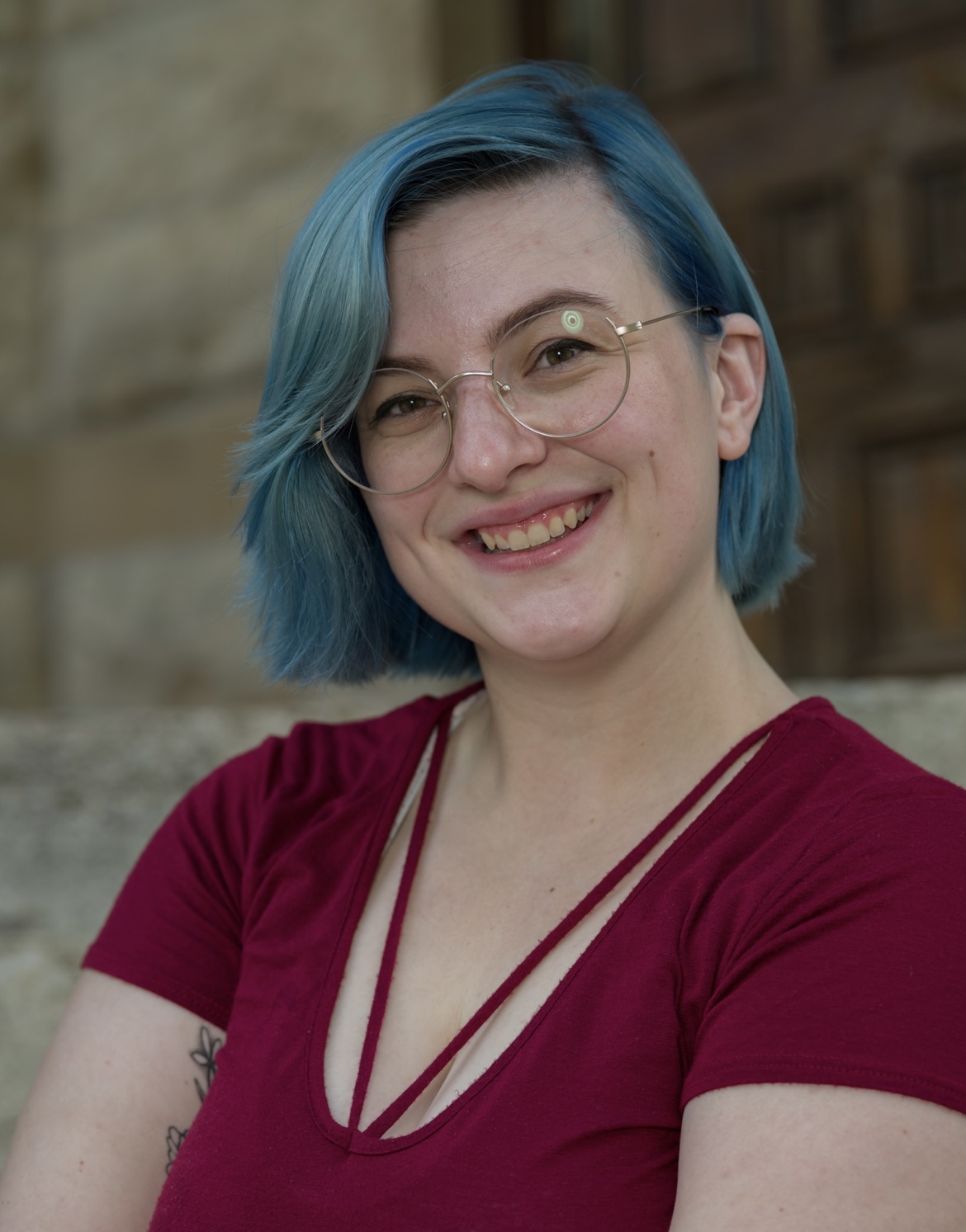 Young woman with blue hair, glasses, and a red top smiling outdoors.