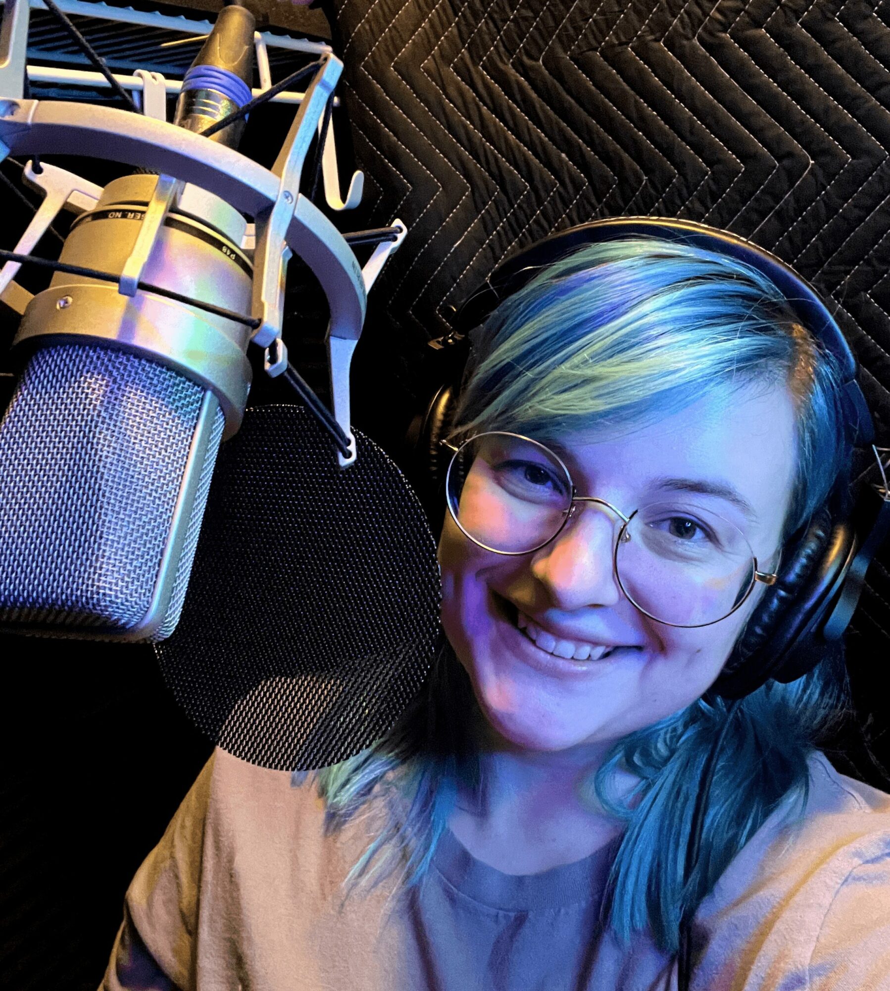 Person with colorful hair and glasses smiling in a recording studio with a microphone and headphones.