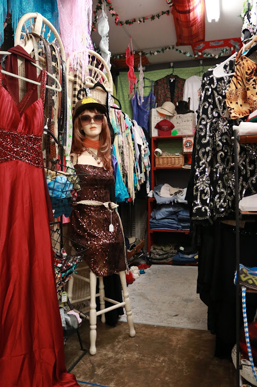 Child wearing sunglasses and a hat, standing among dresses and clothing racks in a store.