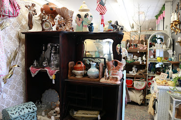 Display of various decorative items and ceramics on shelves in a store, with a background of trees and other furniture.