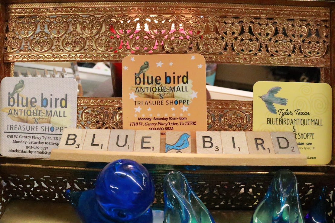 Display of Blue Bird Antique Mall items, including signs and decorative objects, on a shelf with ornate gold trim.