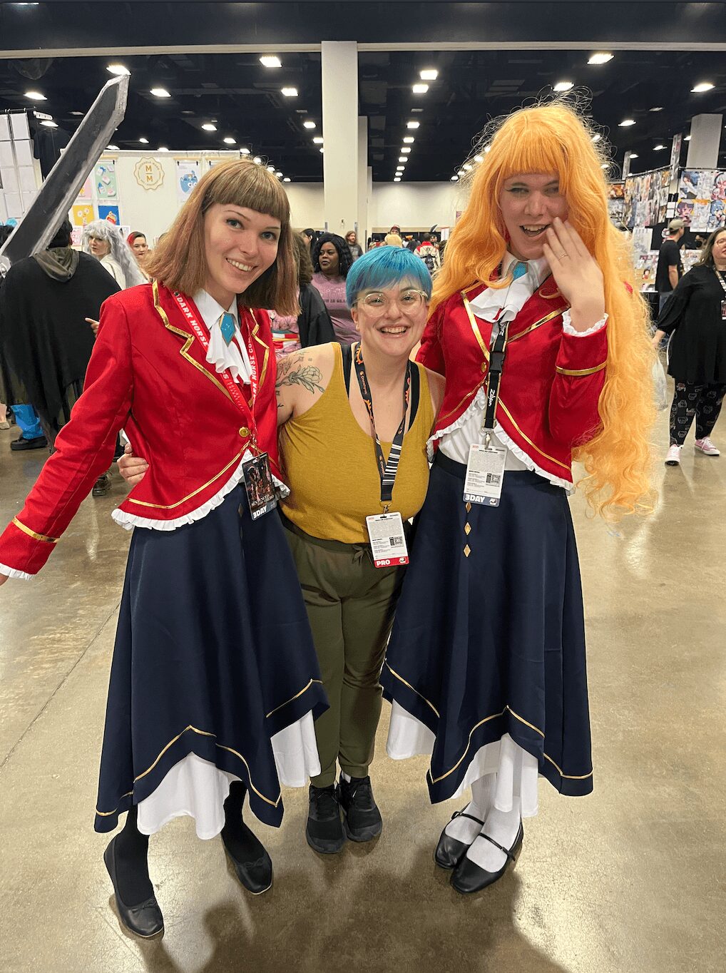 Three people in costumes posing together at an indoor event, smiling, with booths and other attendees in the background.