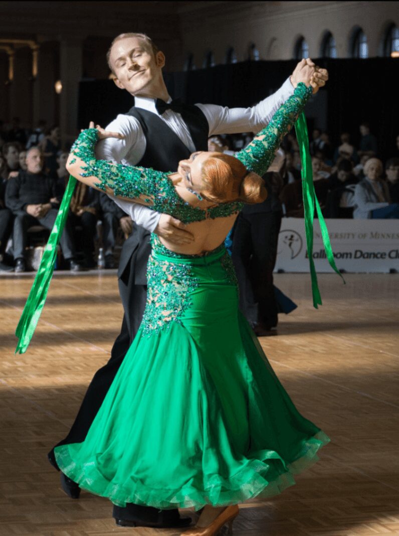 A couple dances ballroom, the woman in a green dress with sequins, the man in a black suit, in a ballroom setting.