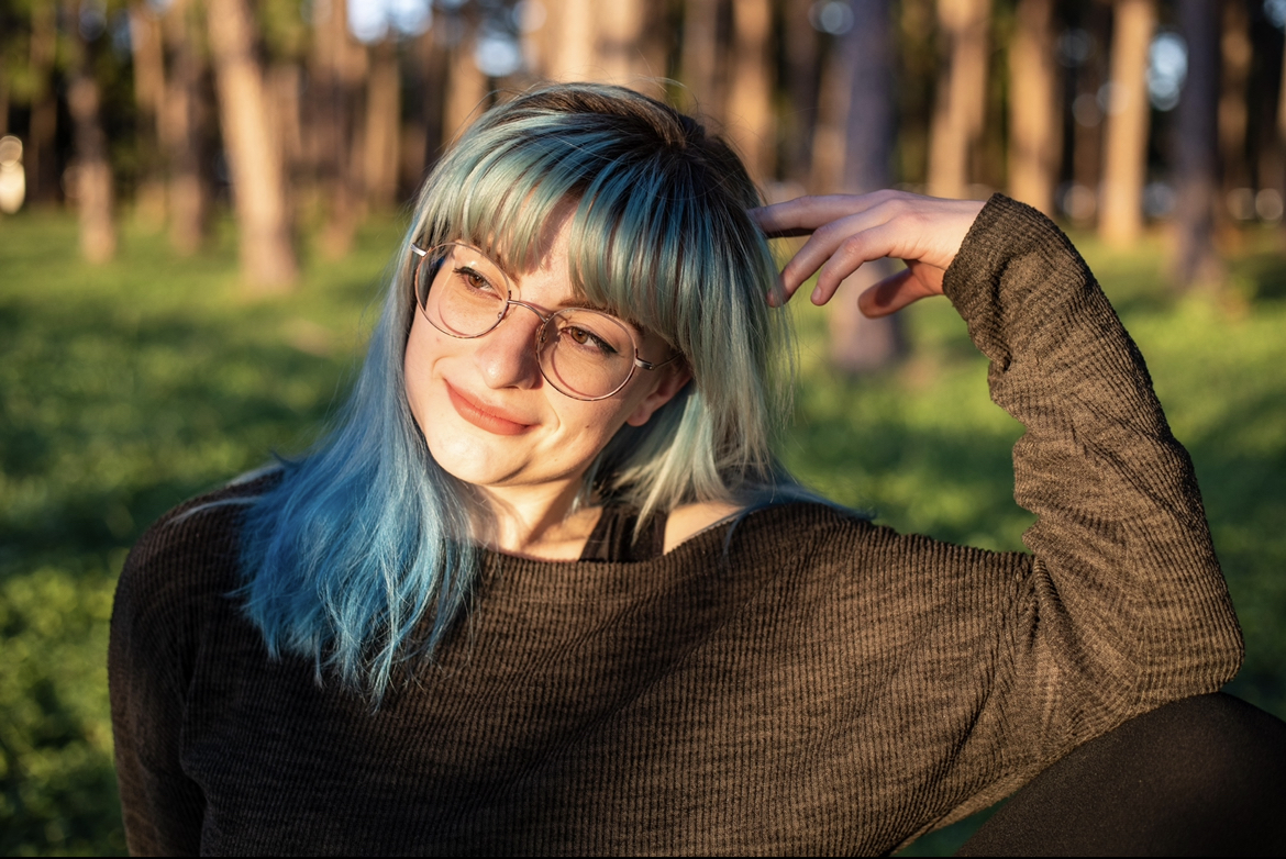 Young woman with blue and green hair, glasses, smiling outdoors in sunlight, wearing a dark sweater, with trees in background.