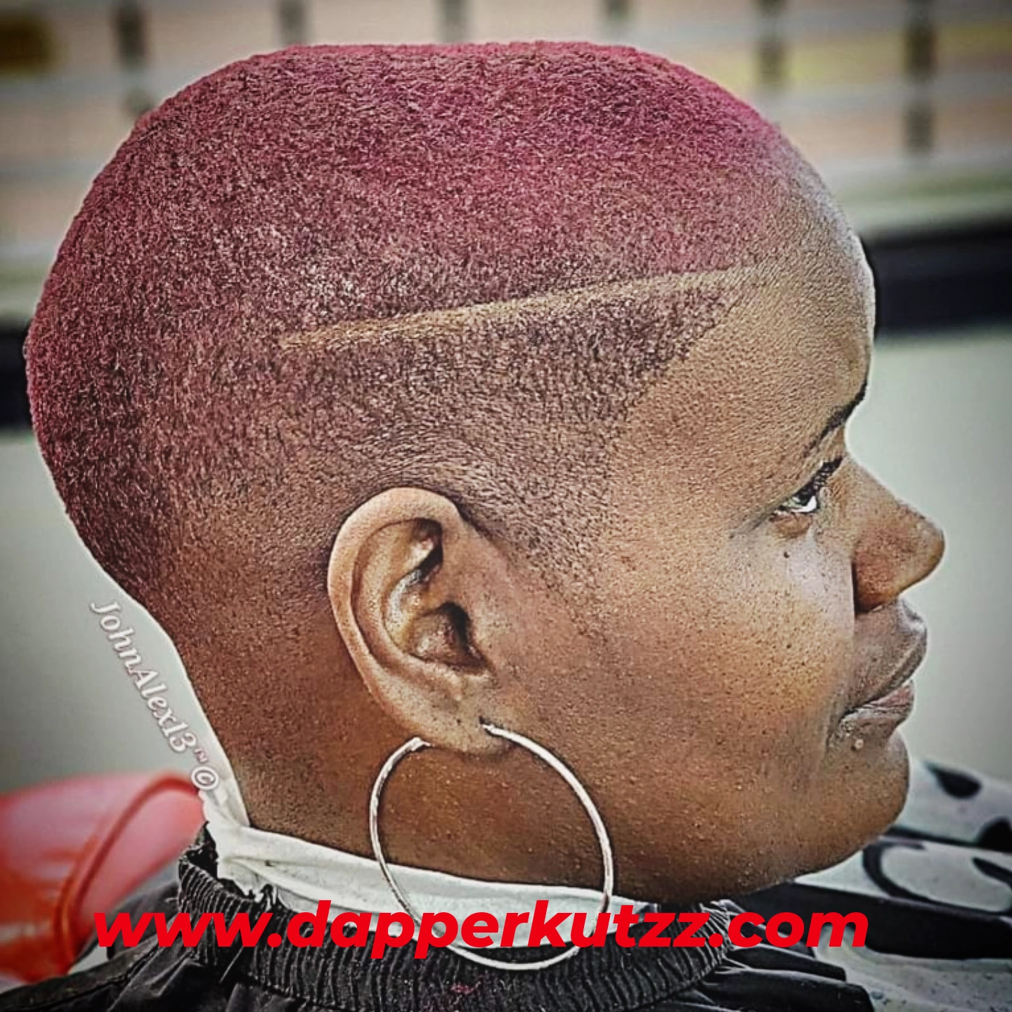 Side profile of a person with a short, pink-colored hairstyle and large hoop earring, sitting indoors.