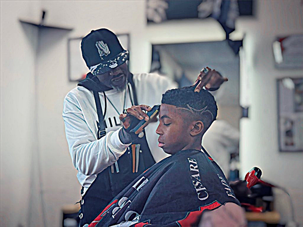 Barber cutting young man's hair in a salon, with shelves and tools in background.