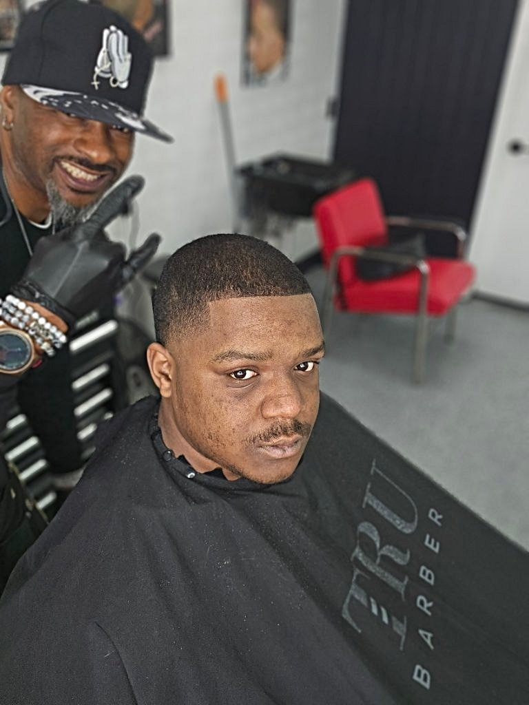A man with a short haircut sits in a barber's chair, looking at the camera, with a barber smiling behind him.