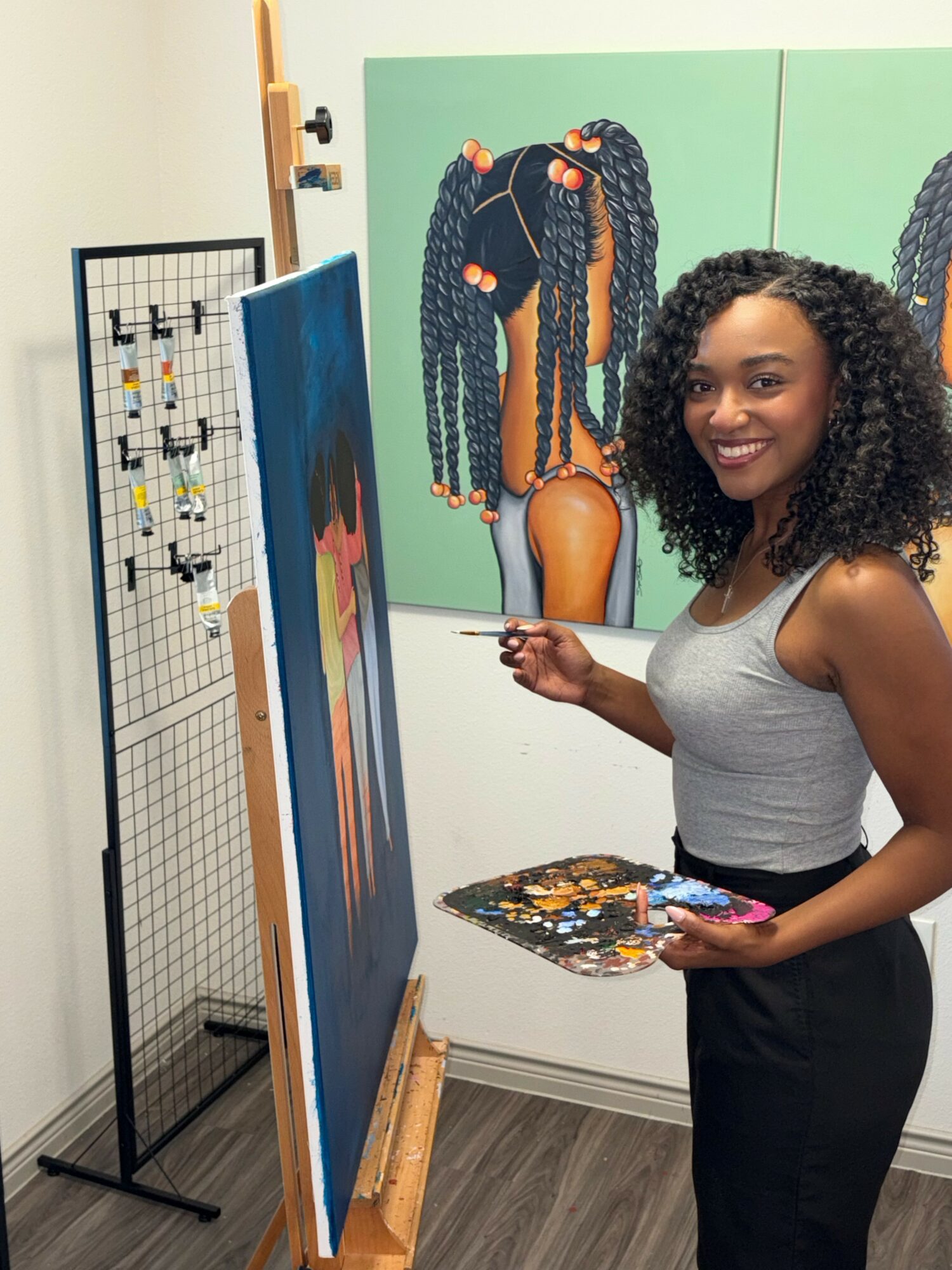 Smiling woman with curly hair holding a paint palette and brush, standing in front of colorful artwork in an art studio.