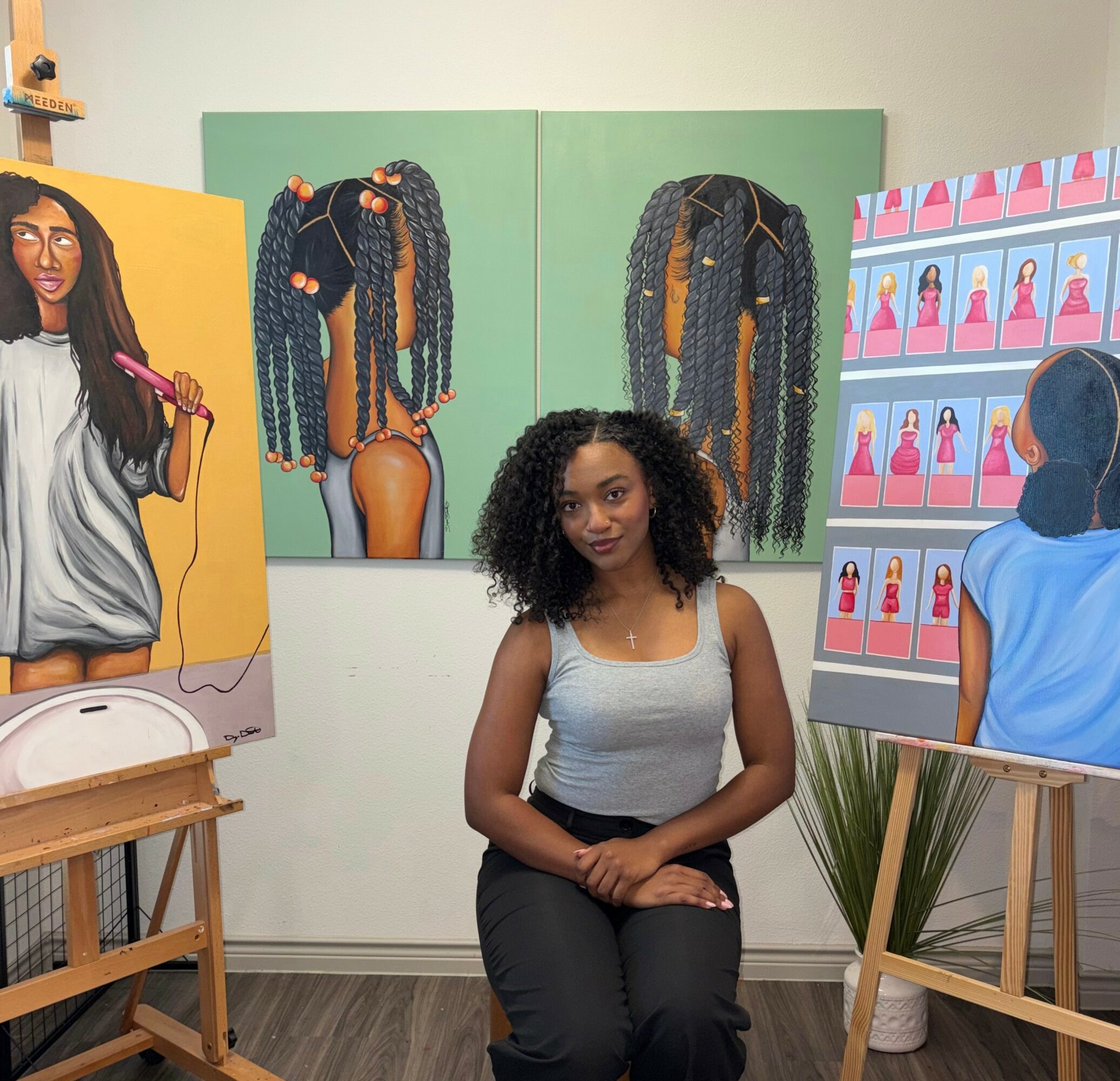 Young woman with curly hair sitting in front of colorful paintings of braided hairstyles, surrounded by art displays.
