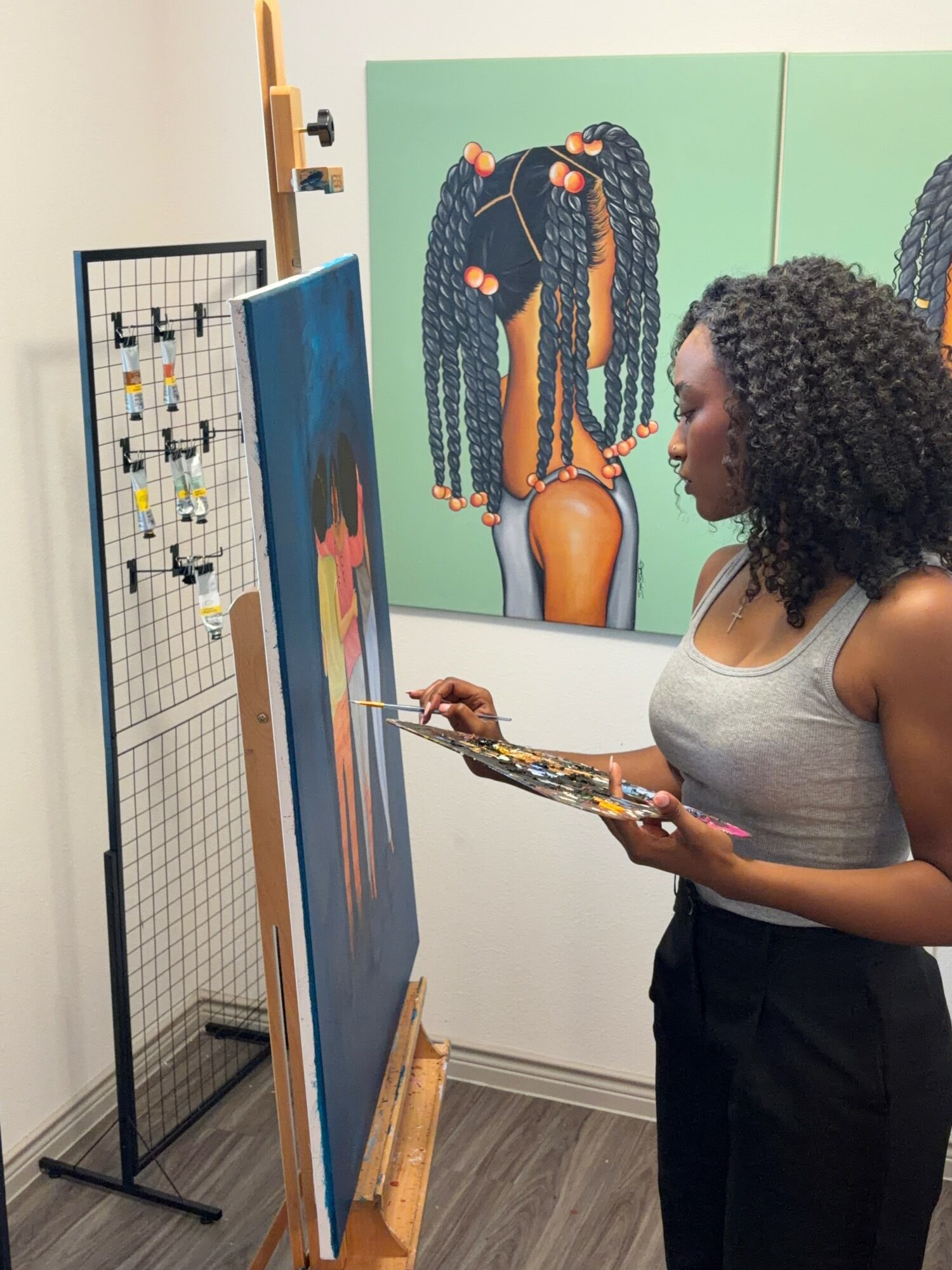 Woman with curly hair painting on canvas in art gallery, colorful artwork behind her, standing on wooden floor.