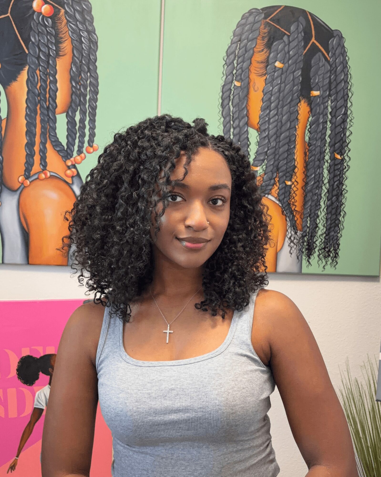 Young woman with curly hair wearing a gray tank top and a cross necklace, standing in front of colorful hair art.