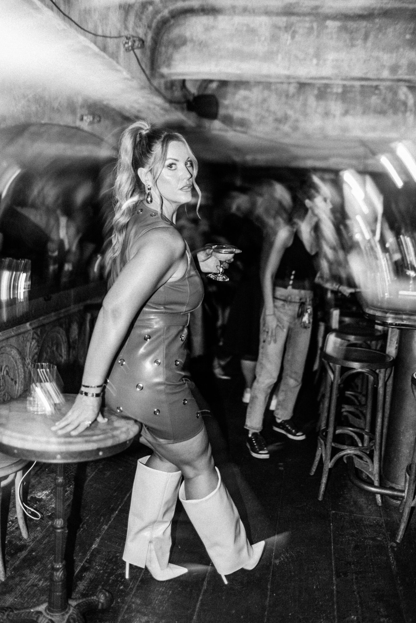 Woman in a dress and boots leaning on a table in a bar with blurred background of people and bar stools.