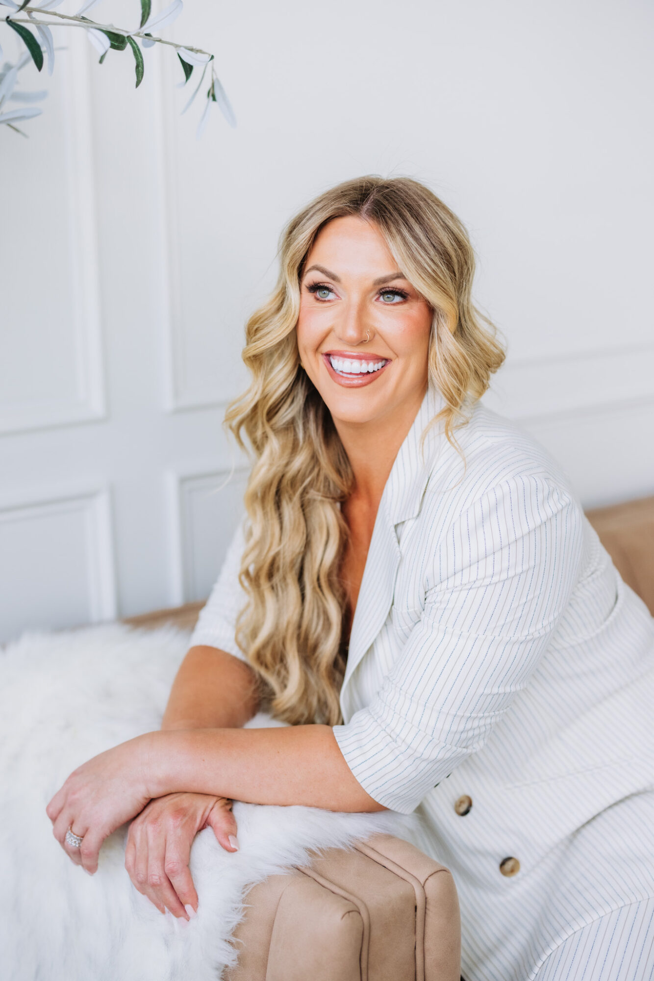 Smiling woman with long wavy blonde hair in a white outfit, sitting on a bed in a bright room.