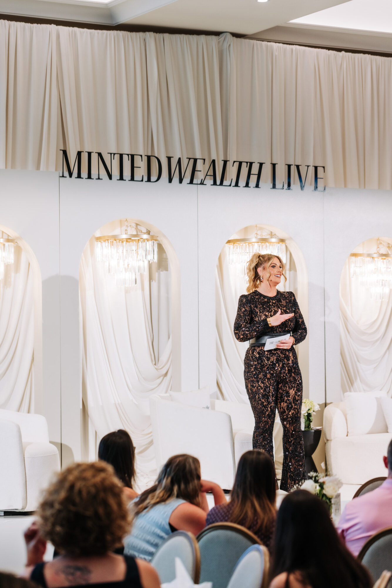 Woman speaking on stage at Minted Wealth Live event, audience seated, white drapes backdrop, event sign above.