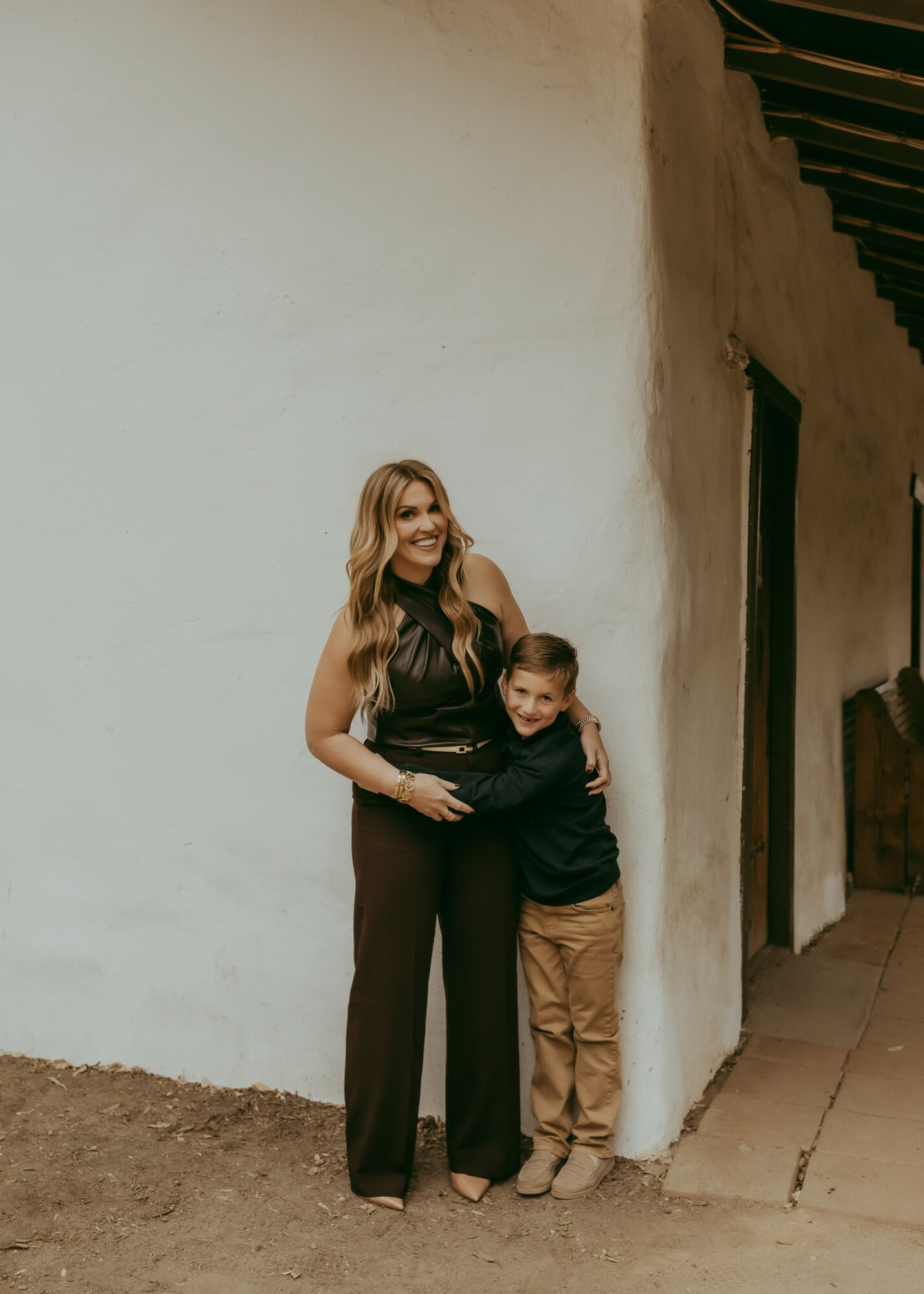 Woman and boy stand against a white wall, smiling, with the woman embracing the boy. They are outdoors.