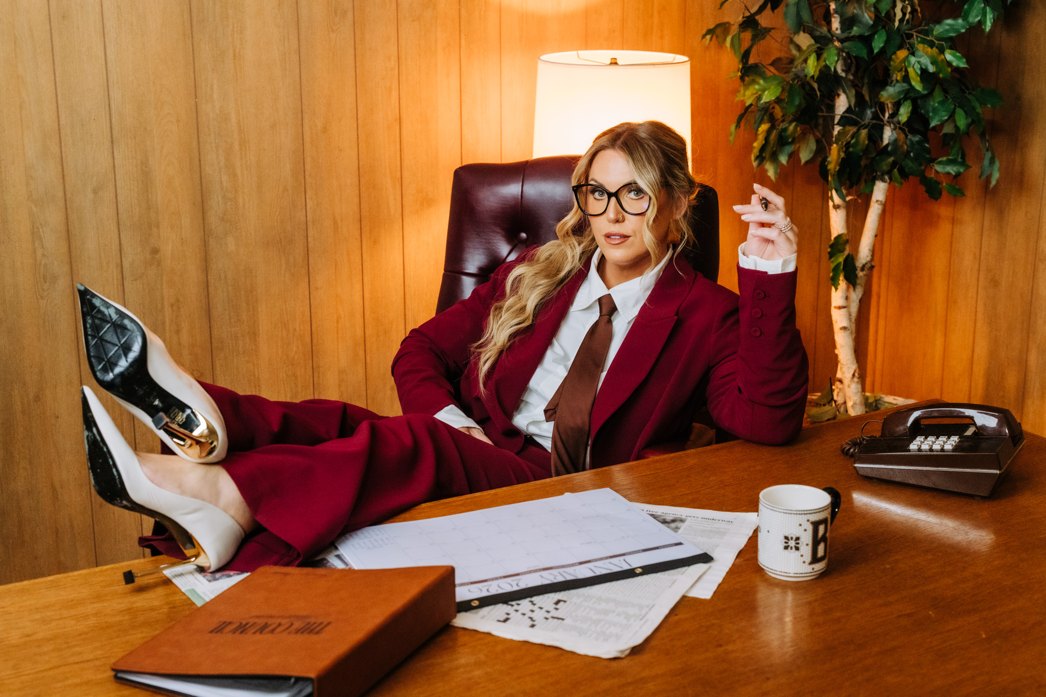 Woman in a burgundy suit and glasses sits at a desk with feet up, holding a phone, in a wood-paneled office.