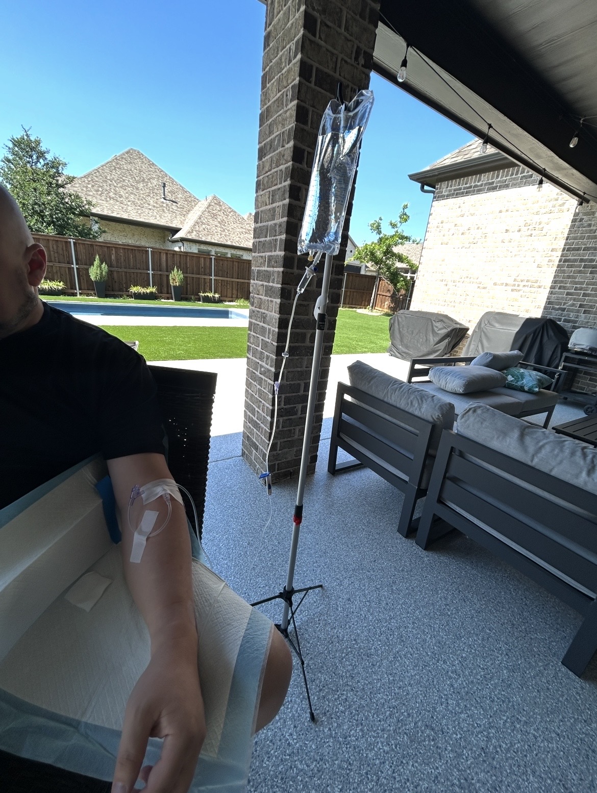 Person receiving IV infusion outdoors on patio with furniture and house in background.