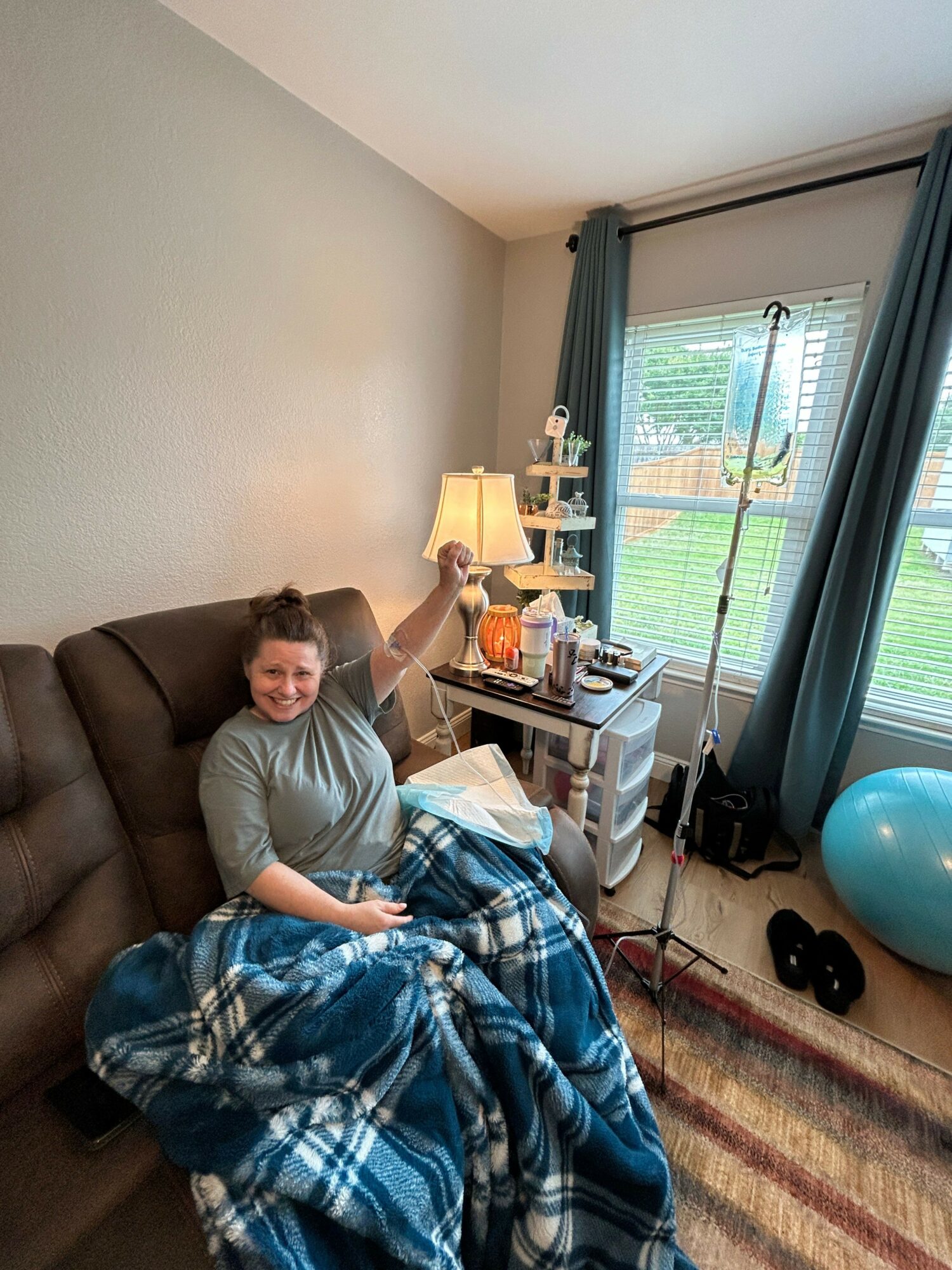Person lying on a couch with a blanket, in a room with a window, curtains, and various items on a table.