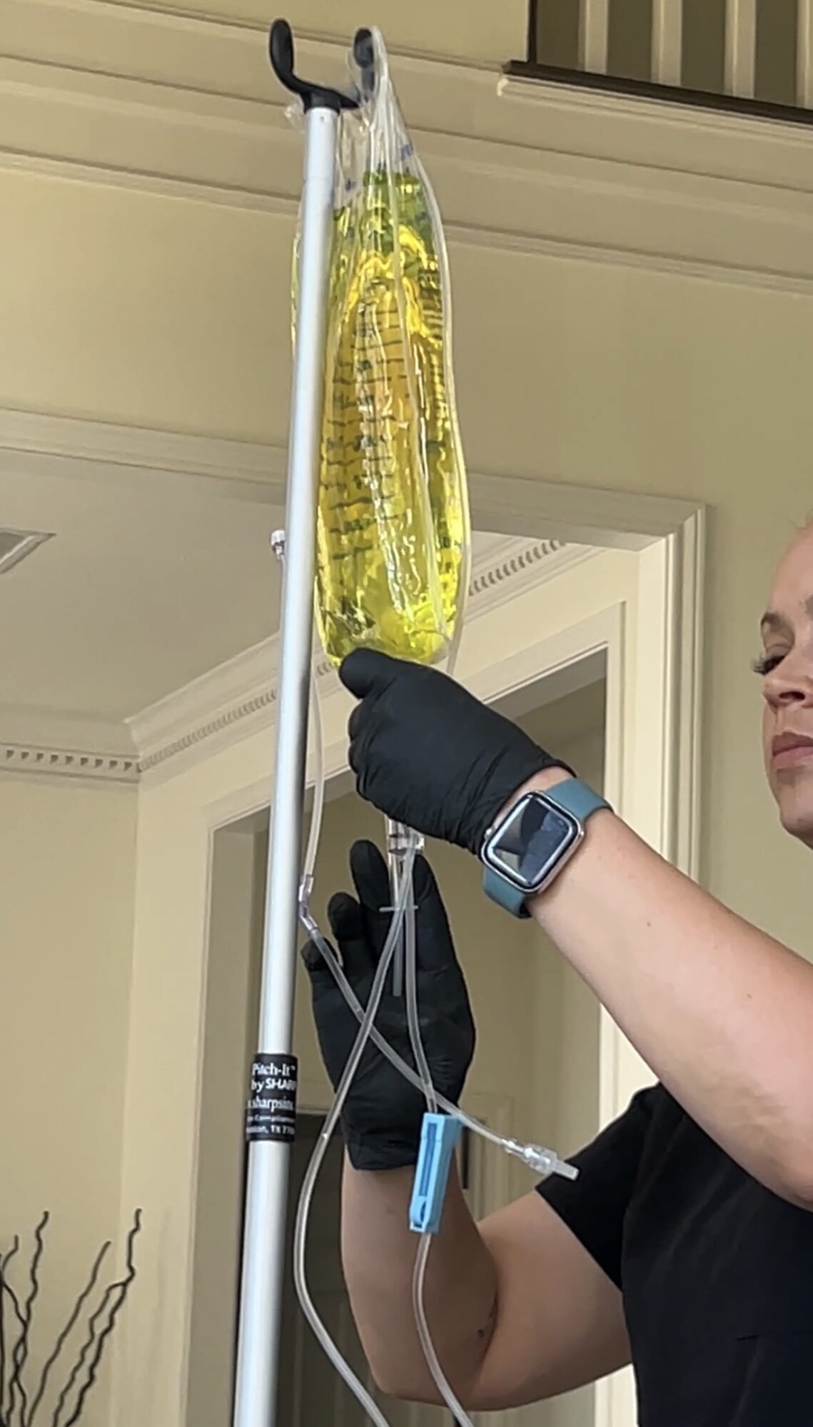 Person in black gloves and watch holds IV bag with yellow liquid, attached to tubing, in a room with cream-colored walls.