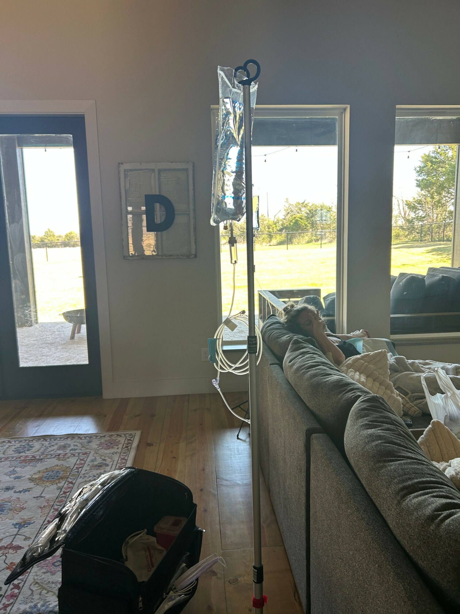 Person sitting on a sofa with an IV drip stand nearby, looking out a large window with a view of trees and outdoor area.