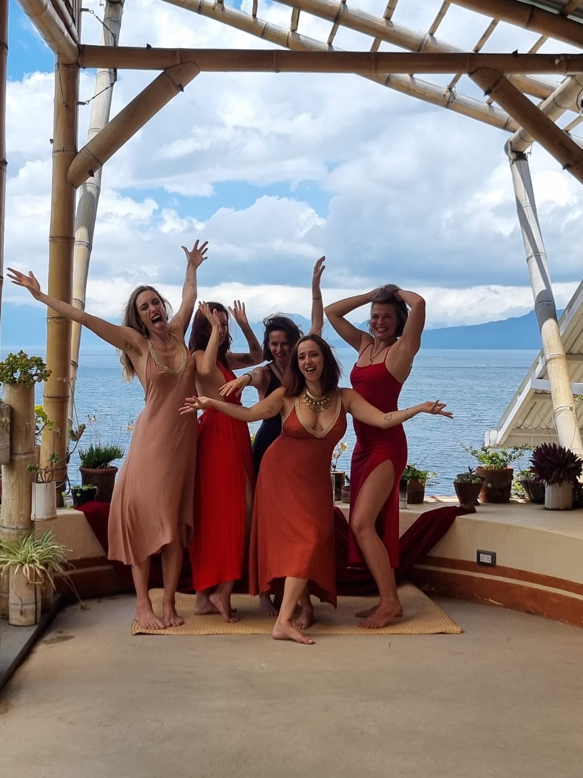 Five women in colorful dresses pose on a wooden platform with a scenic ocean view and cloudy sky in the background.