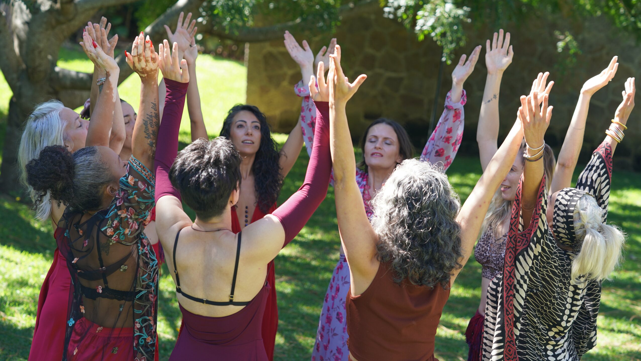 Group of women standing outdoors with arms raised, trees and sunlight in background.