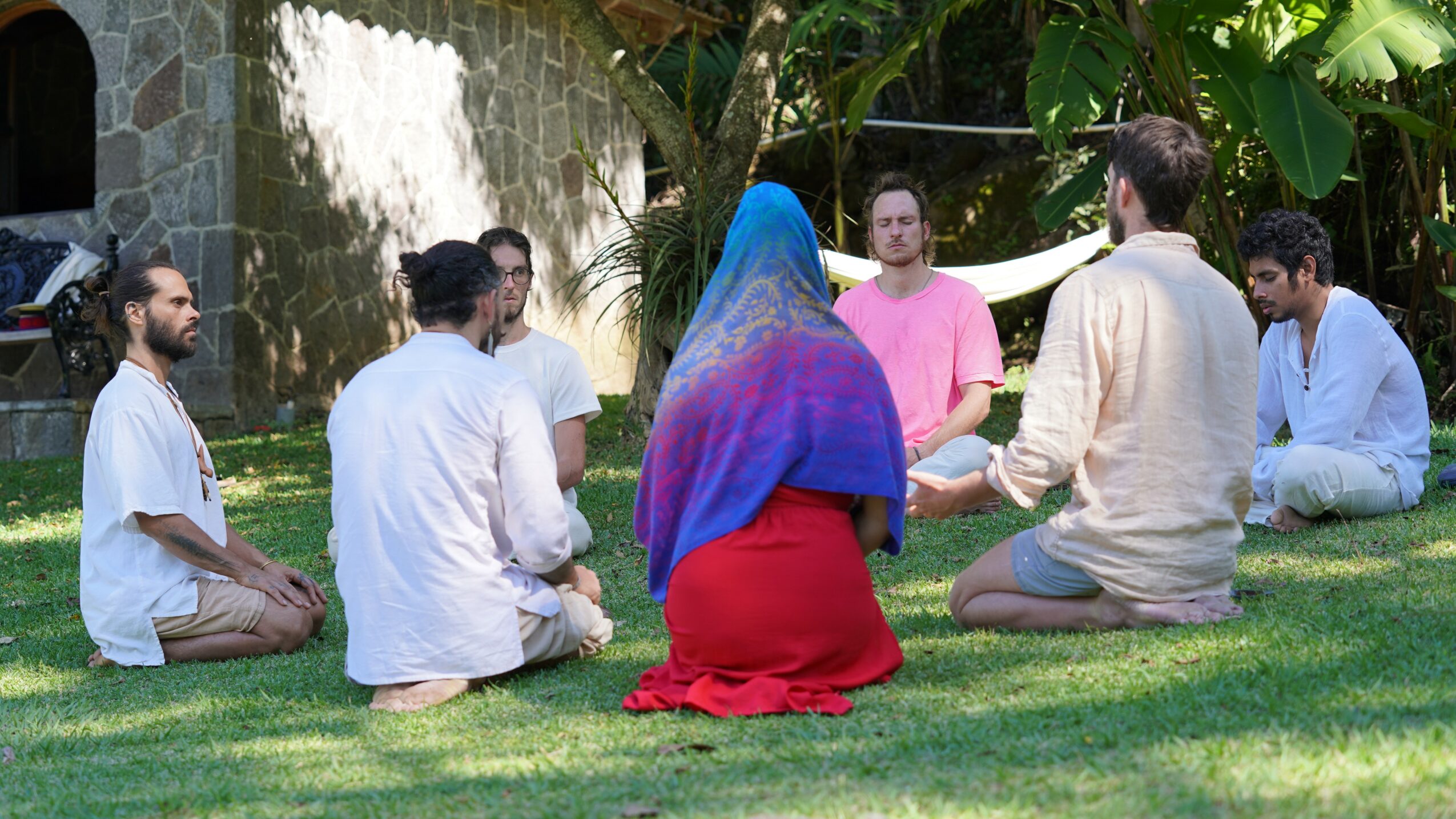 Group of people sitting on grass in a circle outdoors, some with hands folded, surrounded by trees and a stone wall.